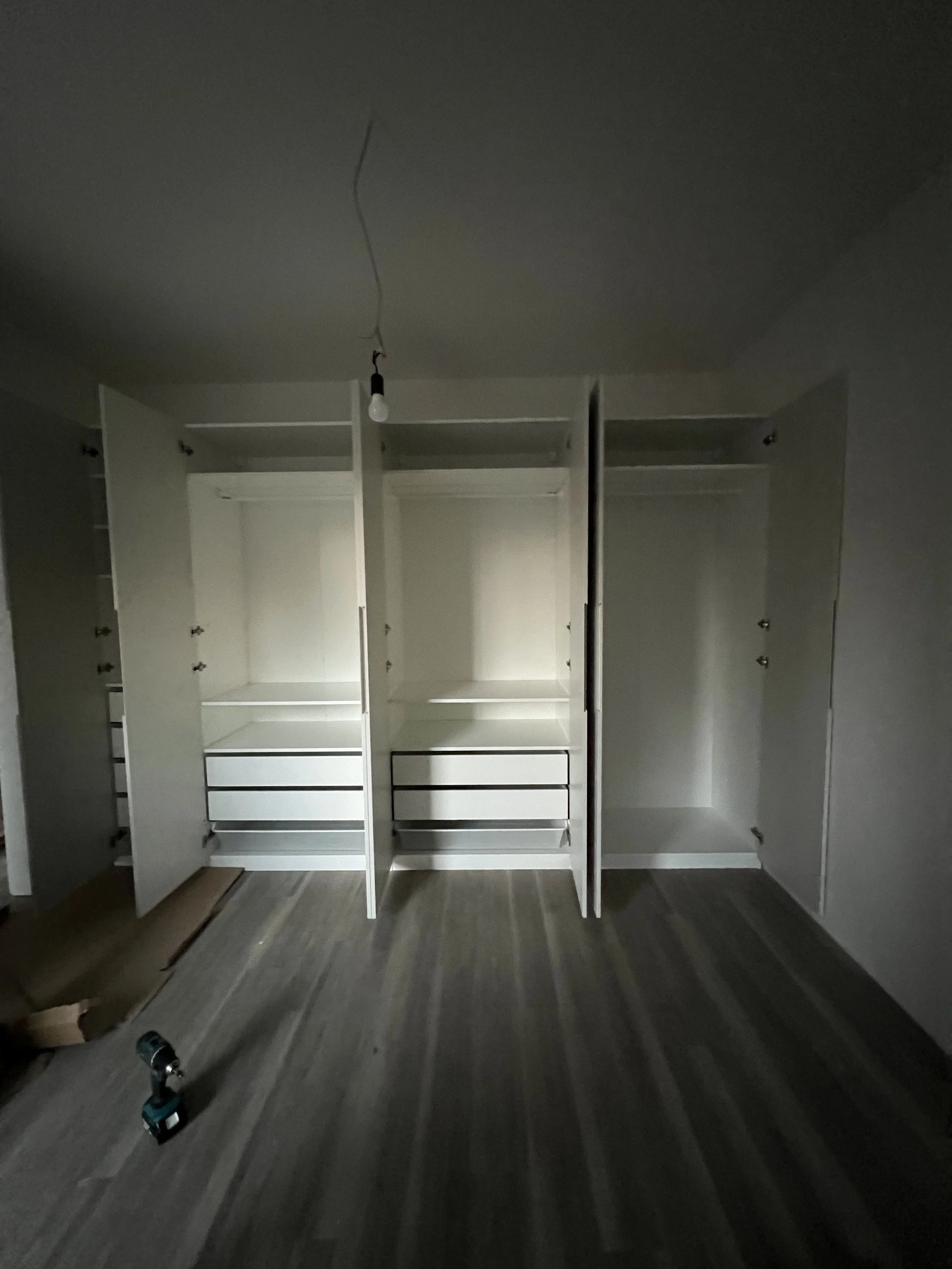 Unbelegter Raum mit weißen Einbauschränken, eine Schraubenzieher liegt auf dem Boden, es ist eine Glühbirne an einer lose hängenden Leitung eingeschaltet.