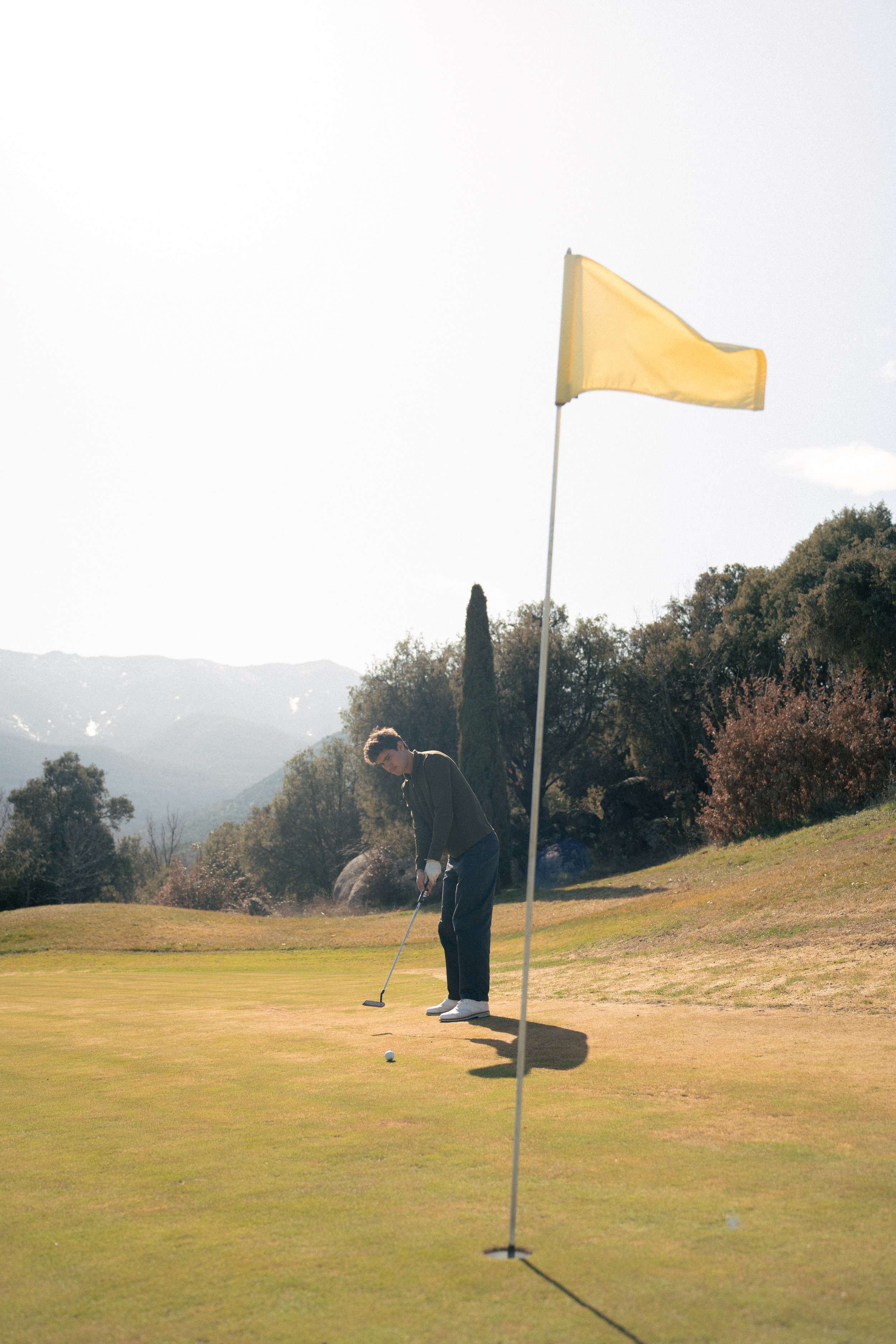 Jugador de golf preparándose para hacer un golpe en el green, cerca de un hoyo con bandera amarilla, en un campo de golf rodeado de árboles y montañas al fondo.