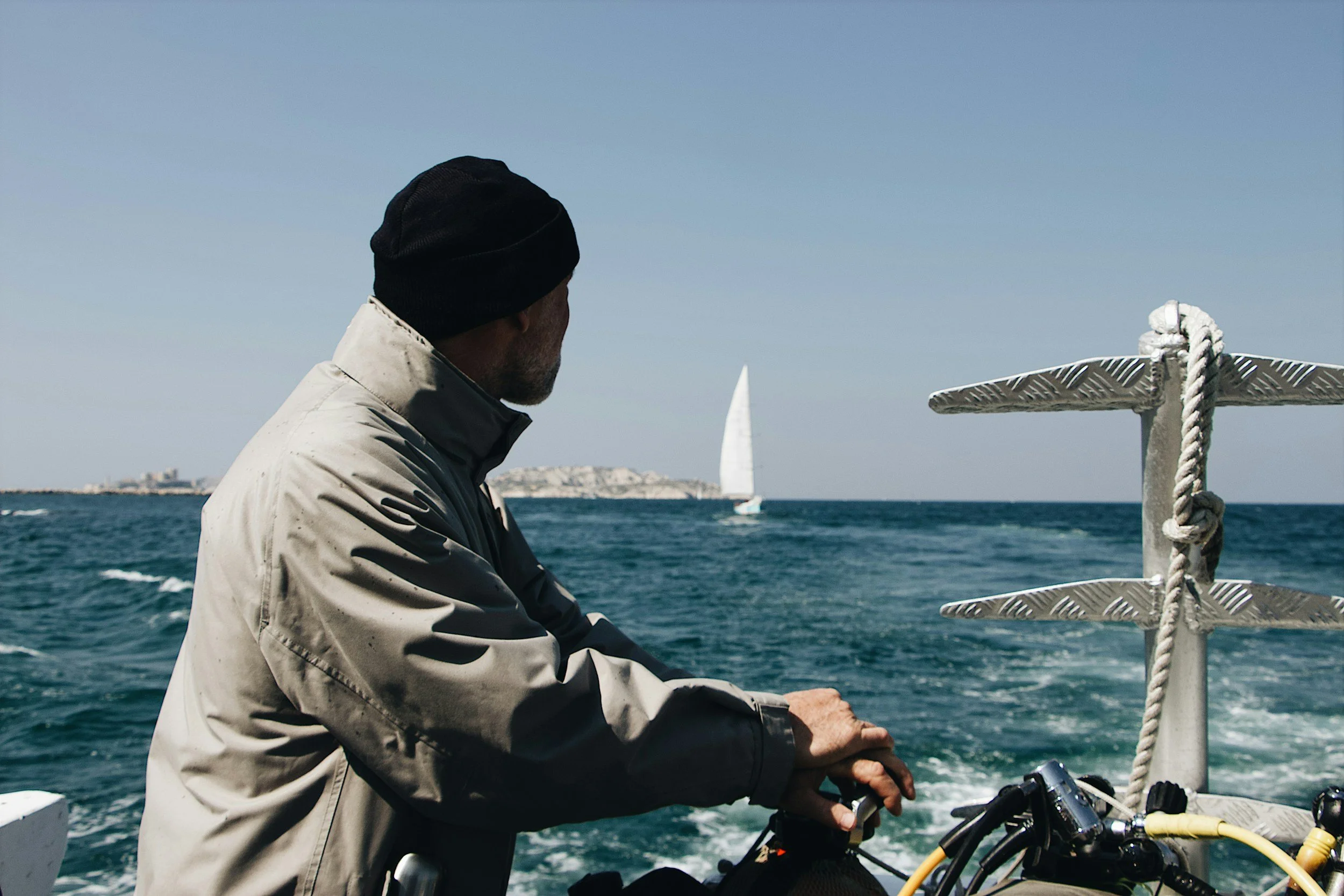 Hombre con abrigo y gorro negro observando el mar desde una embarcación, con un velero en el horizonte y un paisaje costero a lo lejos.