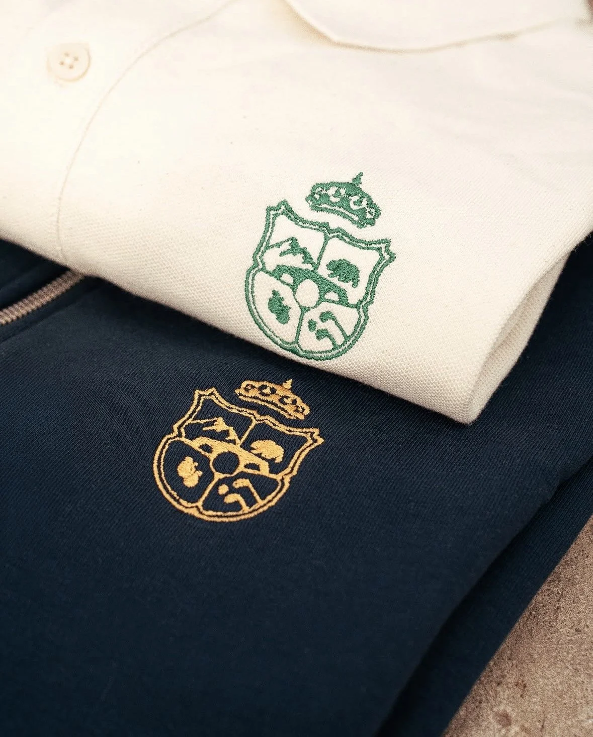 Dos sudaderas apiladas, una blanca con bordado en verde y otra azul con bordado en dorado, ambas con un escudo que tiene diferentes símbolos y una corona en la parte superior.