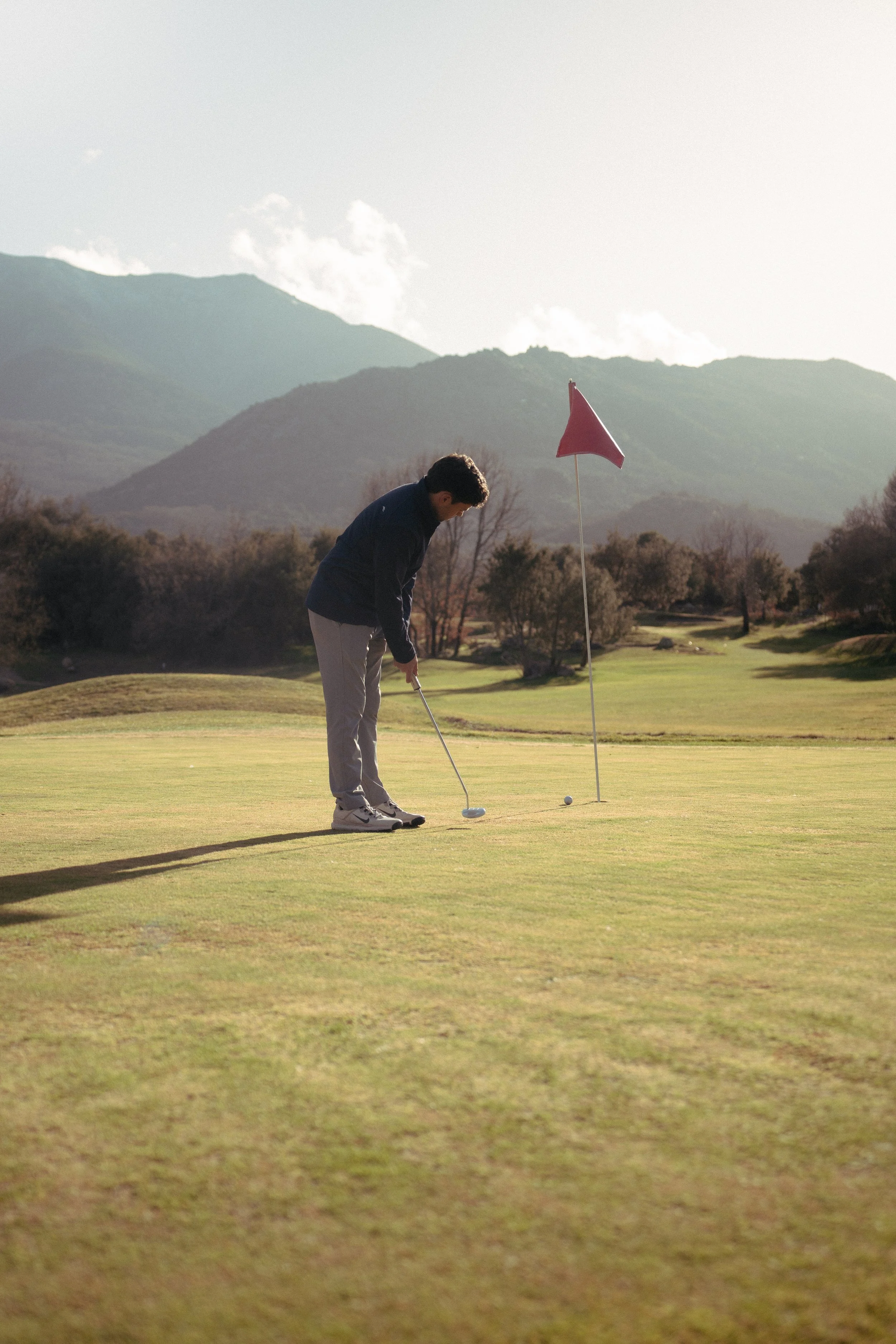 Joven jugador de golf alineando para hacer un golpe en un campo de golf con montaña de fondo y árboles dispersos.