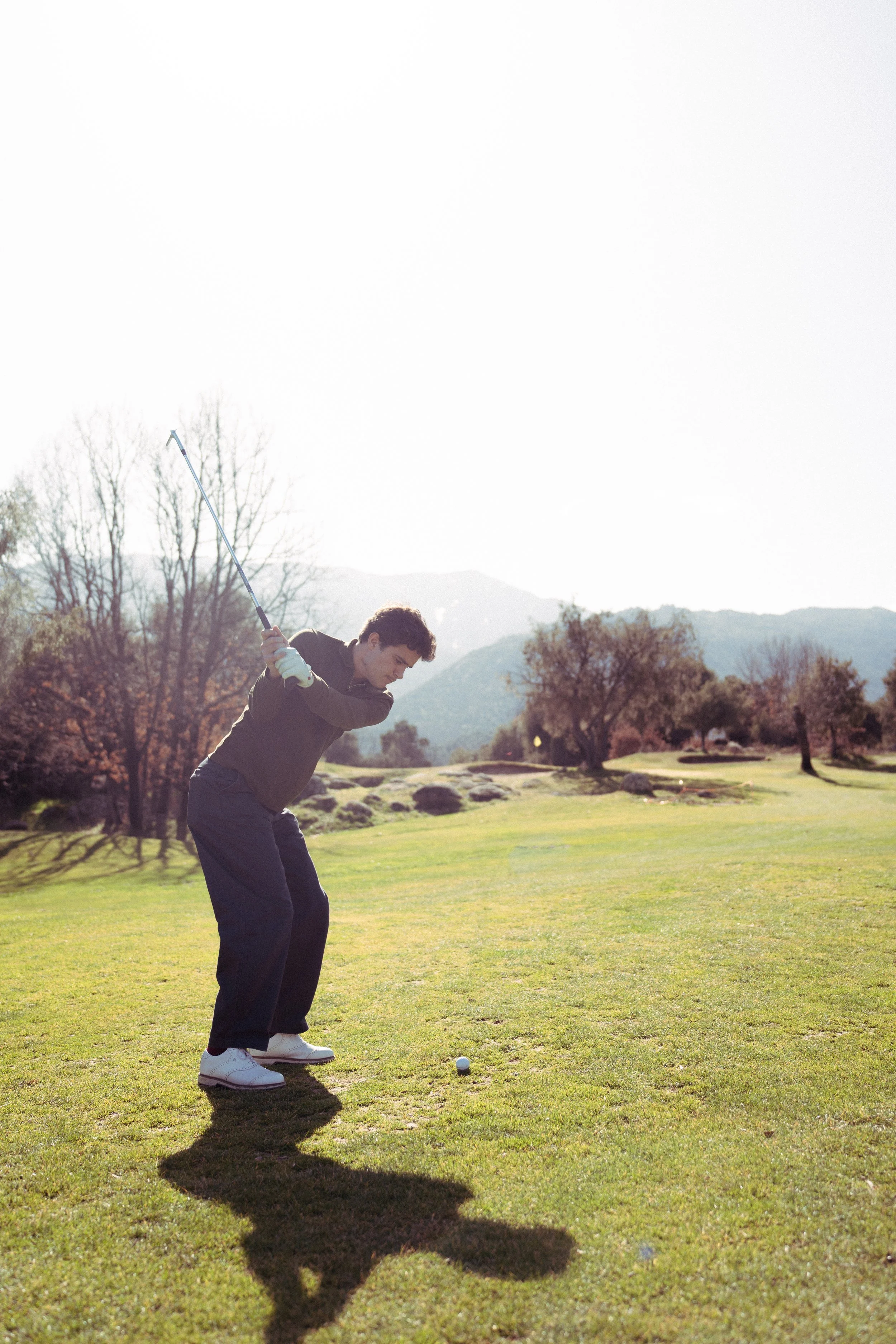 Joven jugando golf en un campo verde con árboles y montañas en el fondo.