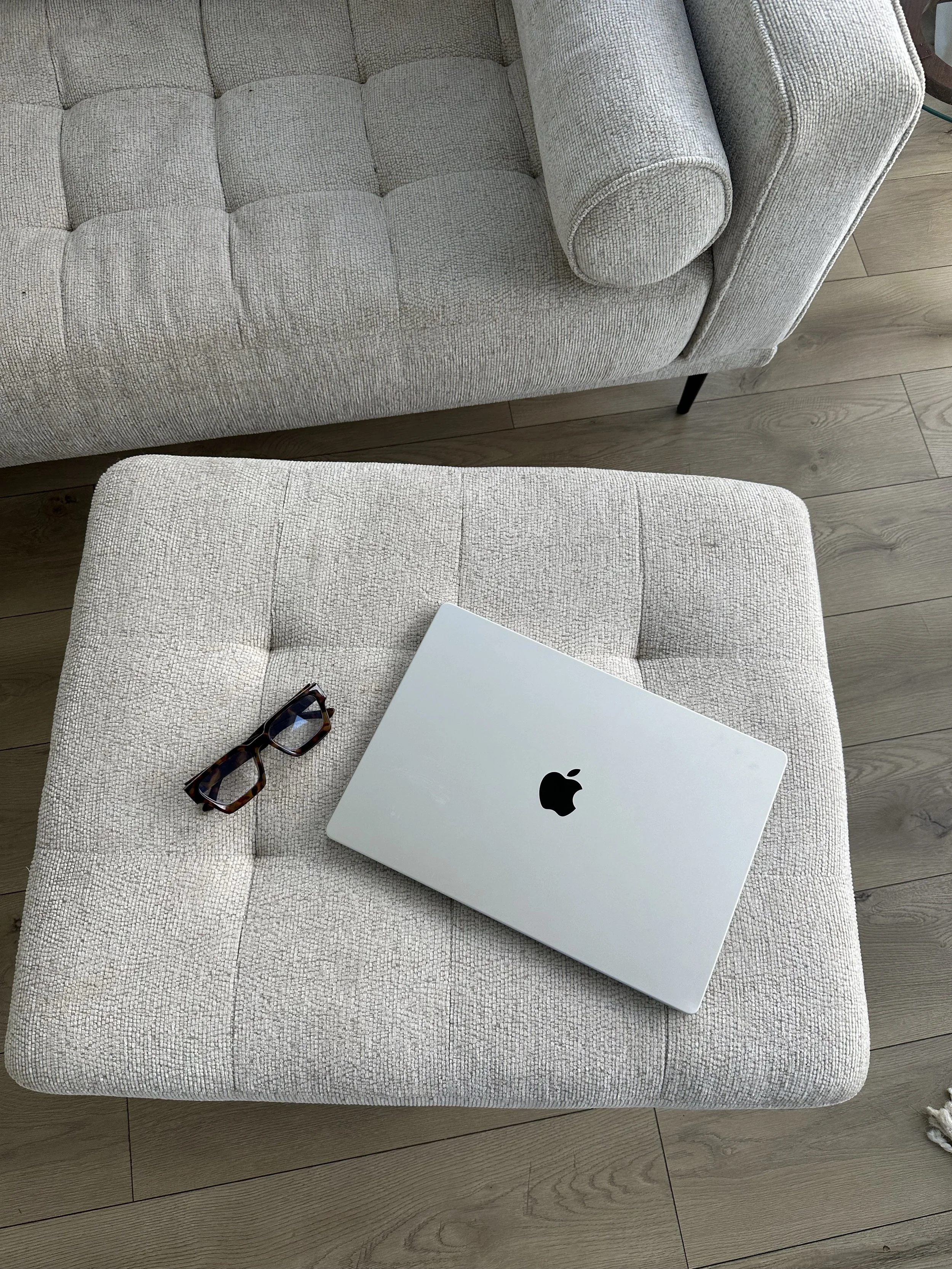 A laptop and a pair of tortoiseshell glasses on a beige, tufted cushioned stool, with a beige sofa in the background on a light wooden floor.