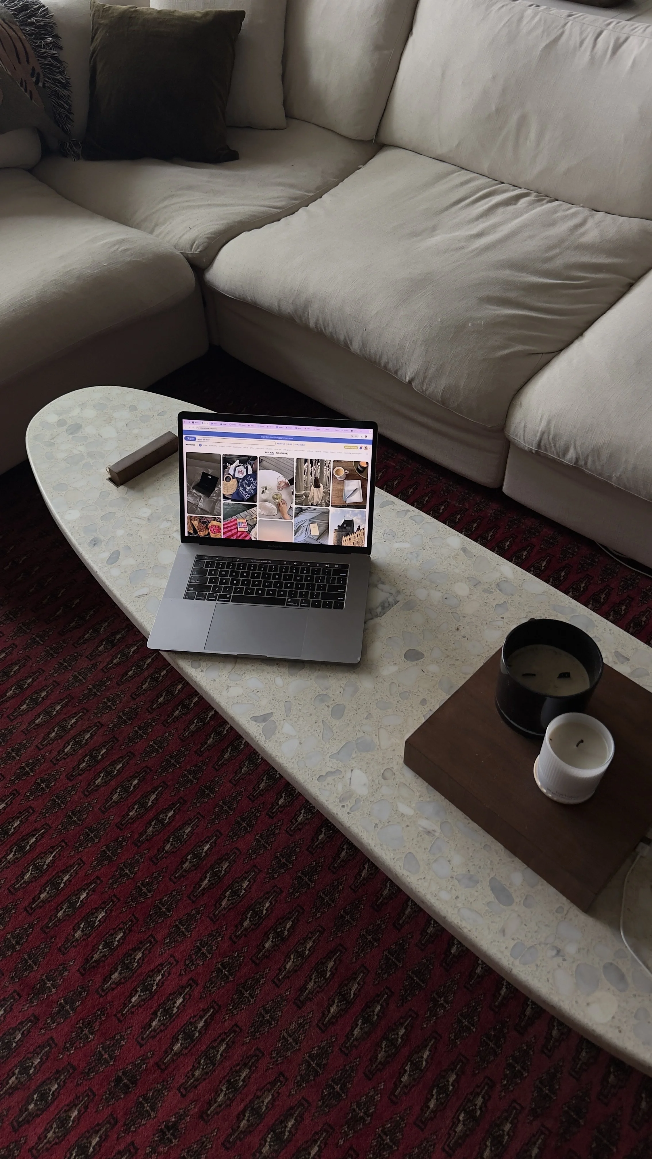 Living room scene with a beige sectional sofa, a coffee table with a laptop displaying a web page, two candles on a wooden tray, and a patterned red carpet.
