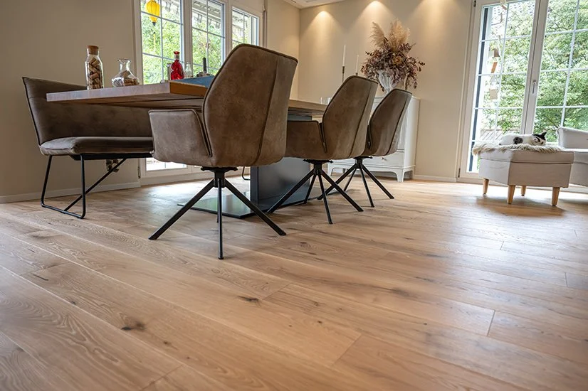 Modernes Esszimmer mit hochwertigem Parkett, Holztisch, sechs braunen Stühlen und großen Fenstern, durch die Natur zu sehen ist. Im Hintergrund steht eine weiße Kommode mit Dekorationen, und auf einem Sessel liegt eine Katze.