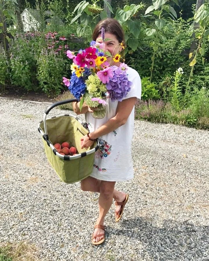 A woman holding a large bouquet of colorful flowers and a shopping basket filled with peaches, standing outdoors on a gravel path with lush green plants and flowers in the background.