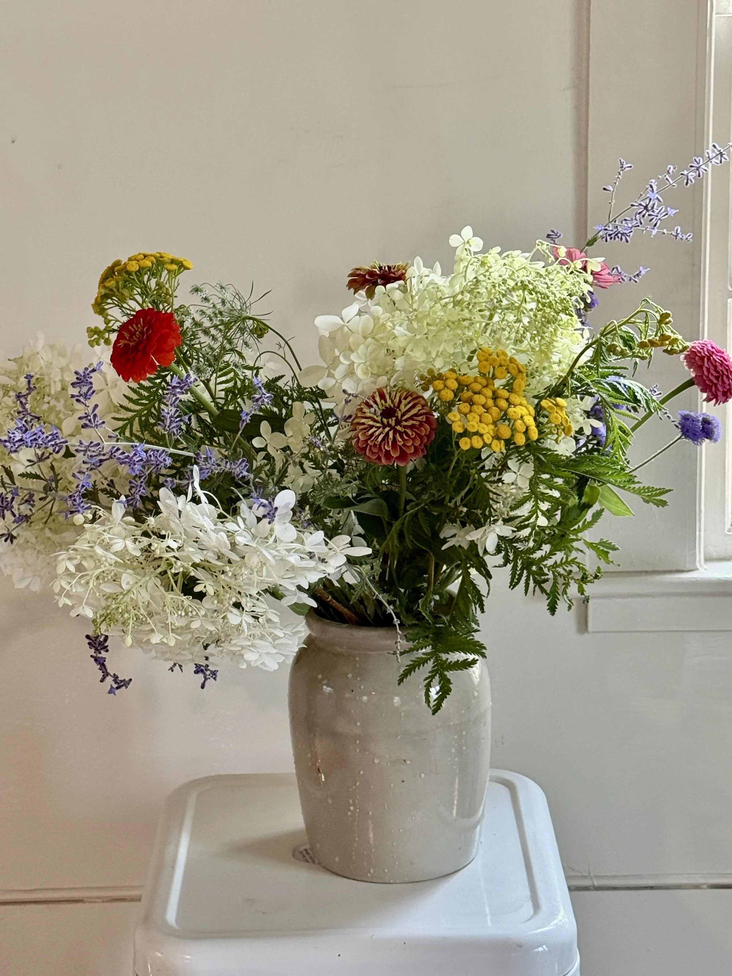 A bouquet of various colorful flowers in a beige ceramic vase on a white table against a blank wall.