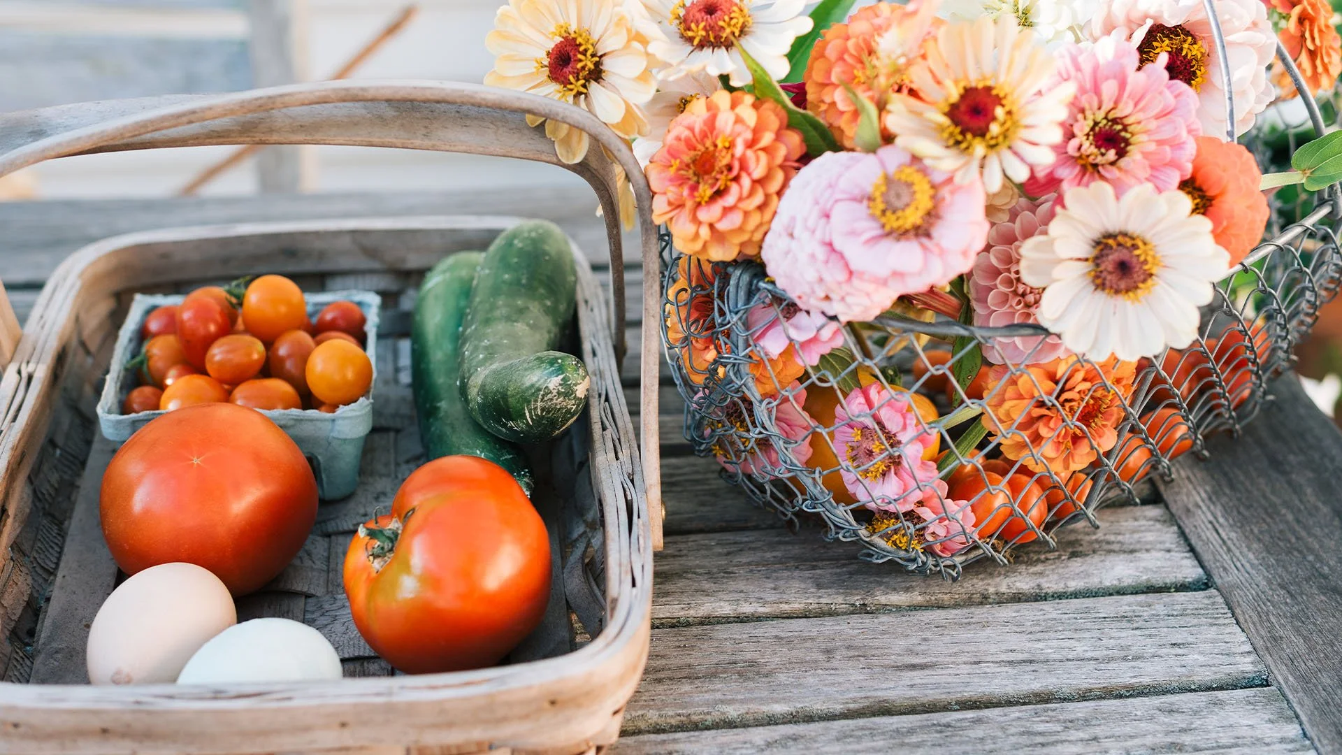 A basket of fresh vegetables, including tomatoes, a cucumber, and eggs, next to a wire basket filled with pink, orange, and white flowers on a wooden surface.