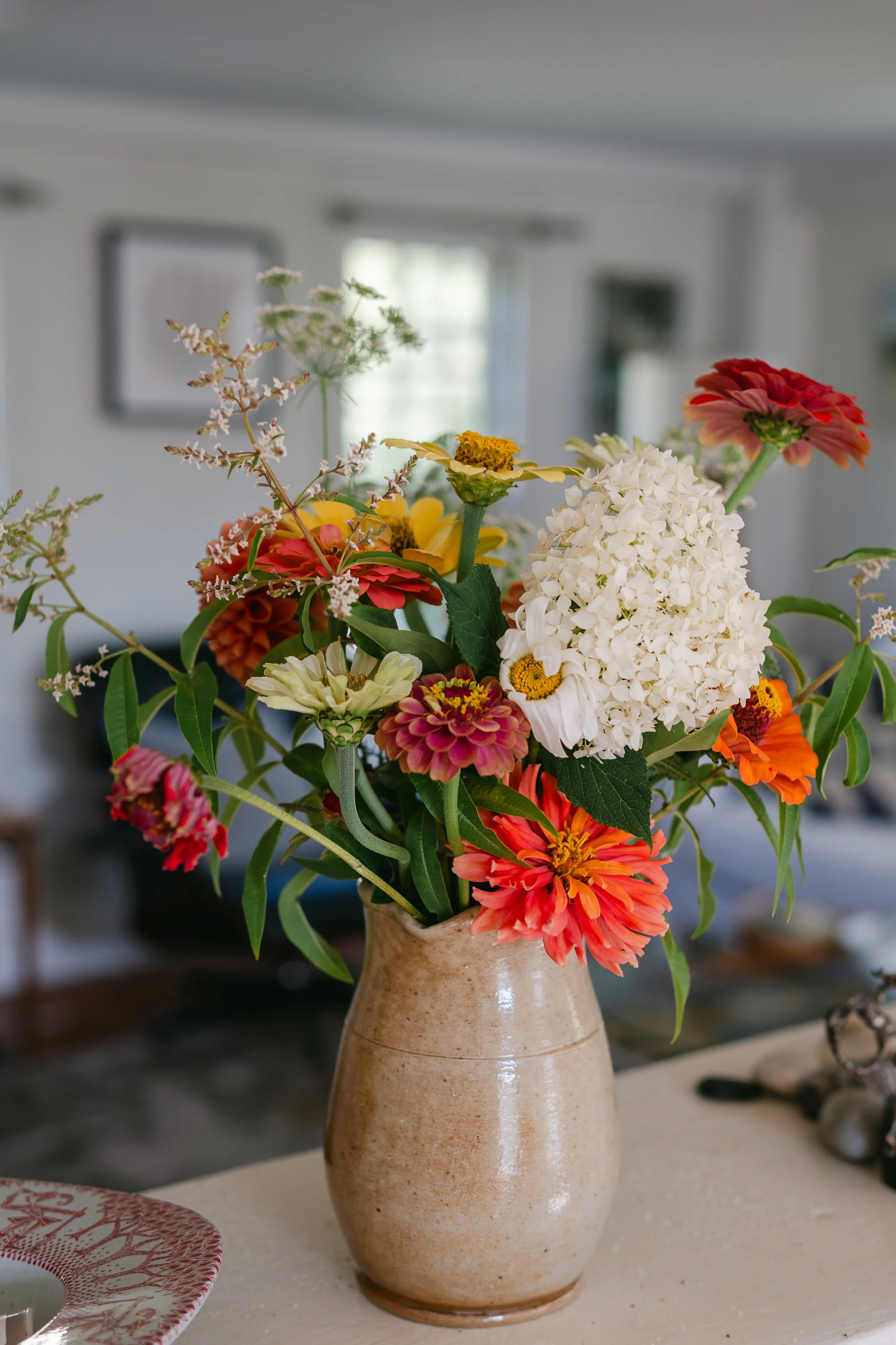 A beige ceramic vase containing a colorful bouquet of assorted flowers including hydrangeas, zinnias, and marigolds, placed on a white table in a living room with blurred background.