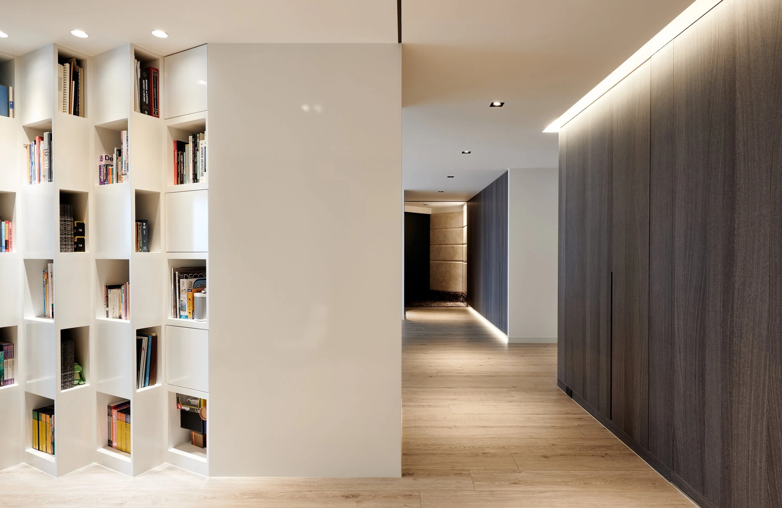 Modern interior hallway with a built-in bookshelf on the left, a white wall in the center, and a dark wood-paneled wall to the right, with recessed lighting on the ceiling and hardwood flooring.