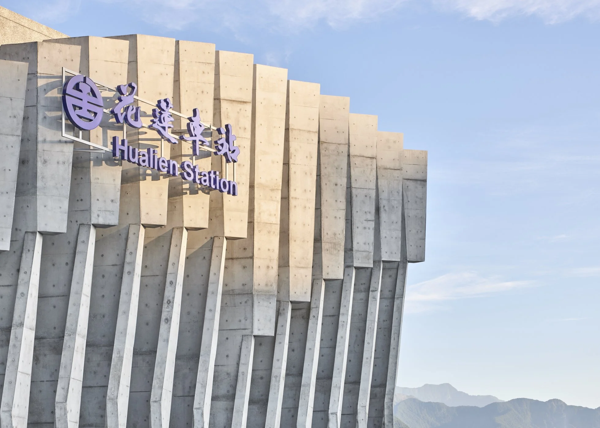 The image shows a modern building with concrete panels and a purple sign that reads 'Hualien Station' in English and Chinese characters, set against a blue sky with some clouds and distant mountains in the background.