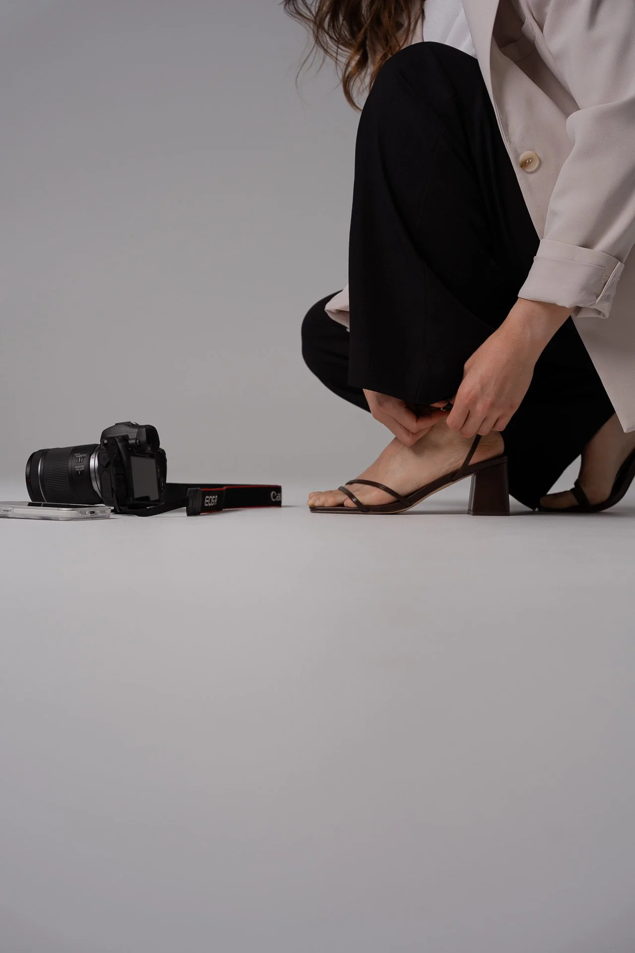 A woman in black pants and a cream blazer is kneeling and adjusting her heel while a camera, phone and camera accessories are placed on the floor nearby.