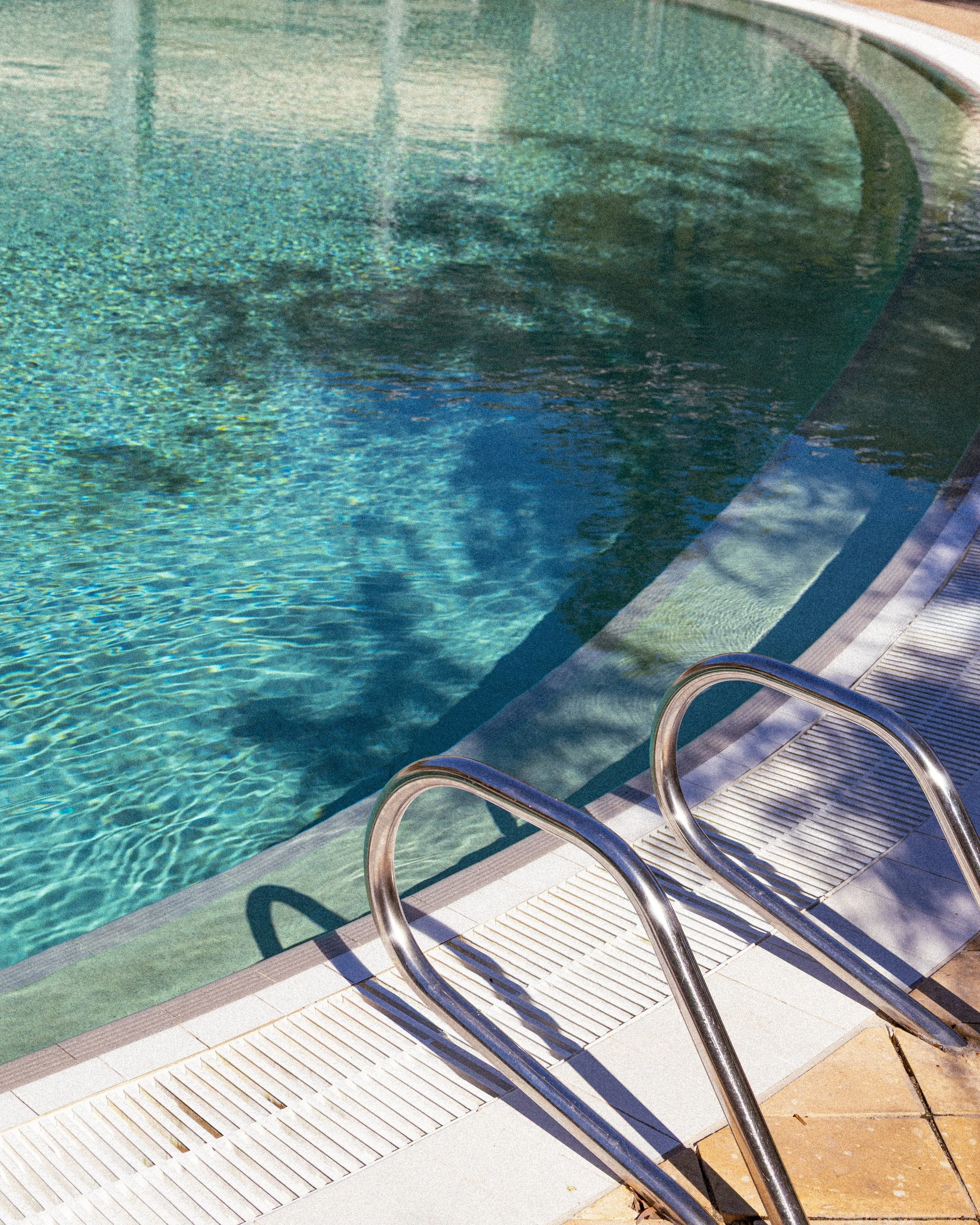 Close-up of a swimming pool with metal handrails on the side, water in the pool, and a white plastic drain cover along the pool edge, with shadows cast on the ground.