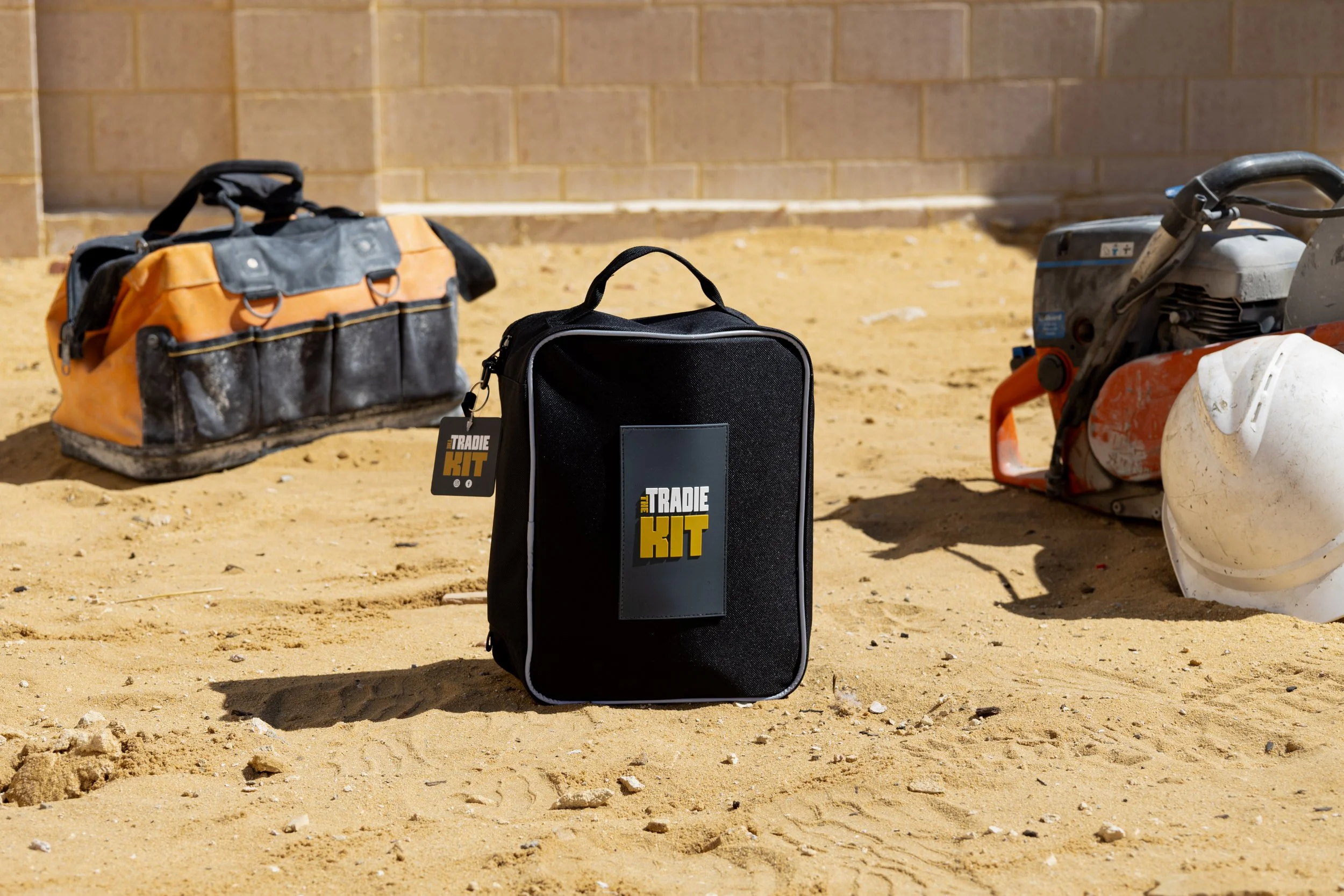Construction tools and equipment on sandy ground including a black toolbox with "TRADIE KIT" label, an orange and black bag, a concrete saw, and a white plastic container.