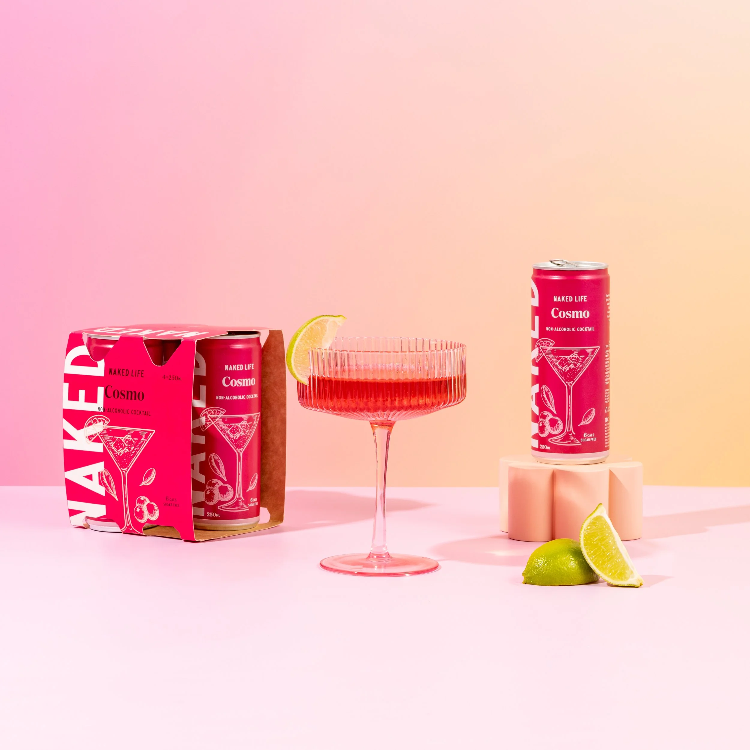 Pink background with two cans of Naked Life Cosmo non-alcoholic cocktail, a pink glass with a lemon wedge, and a pink box with lemon slices and limes.