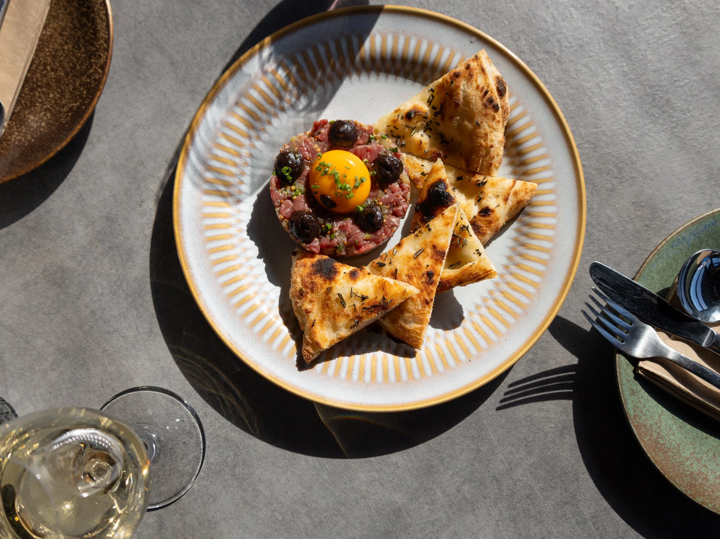 A white plate with a yellow rim containing slices of flatbread, a beef tartare topped with a raw egg yolk, sprinkled with chopped herbs and spices. Part of a glass of white wine and several other dishes are visible around the plate on a grey table.