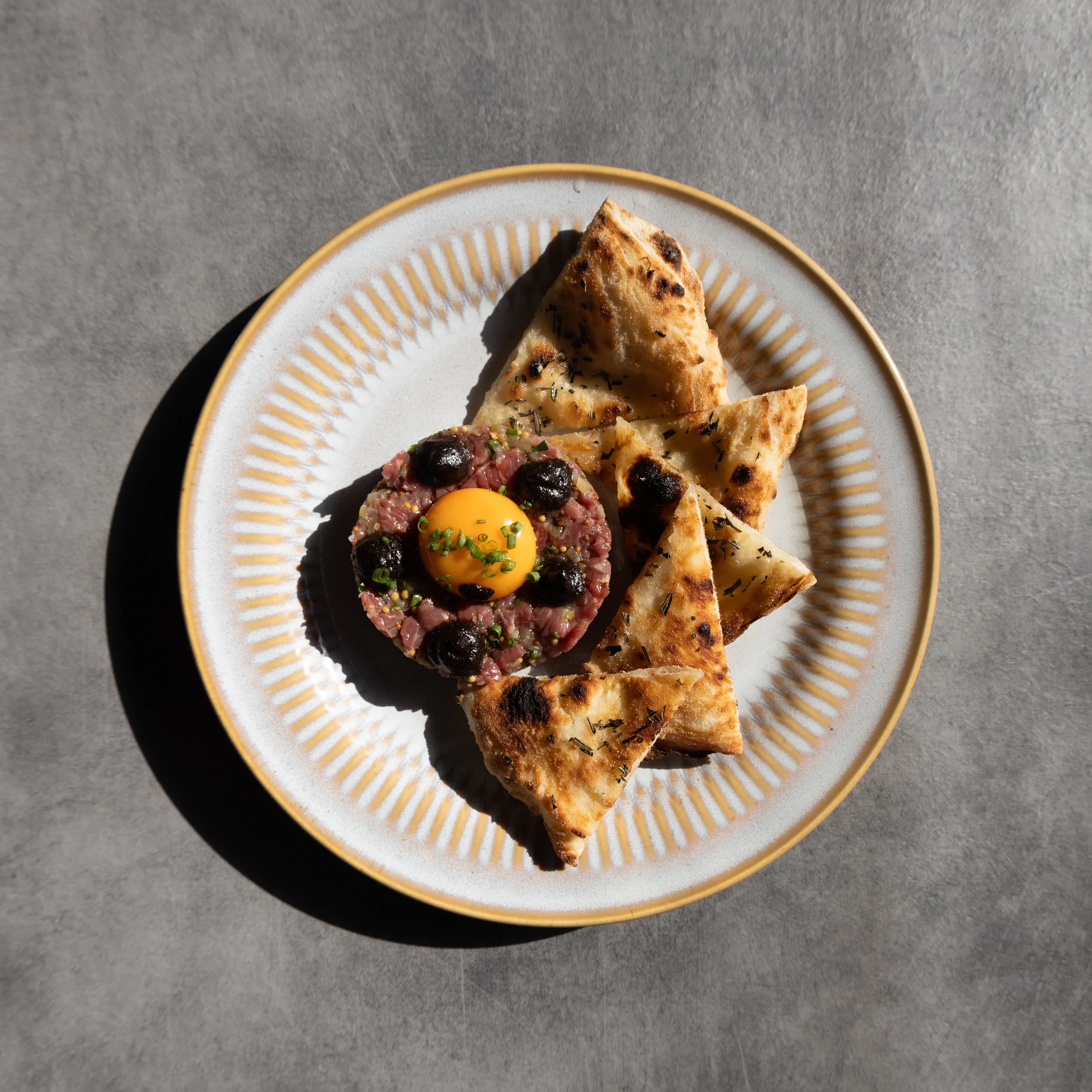 Close-up of a white plate with a gold rim containing flatbread and a meat dish topped with a raw egg yolk and herbs, on a gray surface.