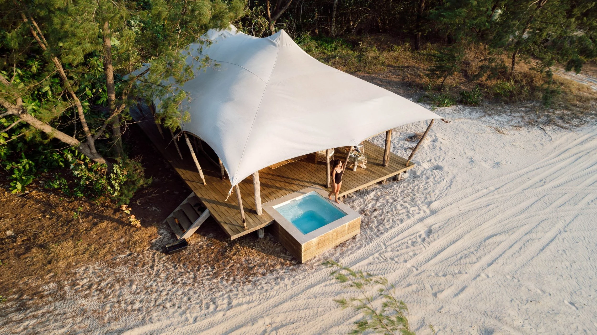 An aerial view of a beachside tent with a white canopy, wooden deck, and a small pool, surrounded by trees and sandy beach, with a woman standing on the deck.