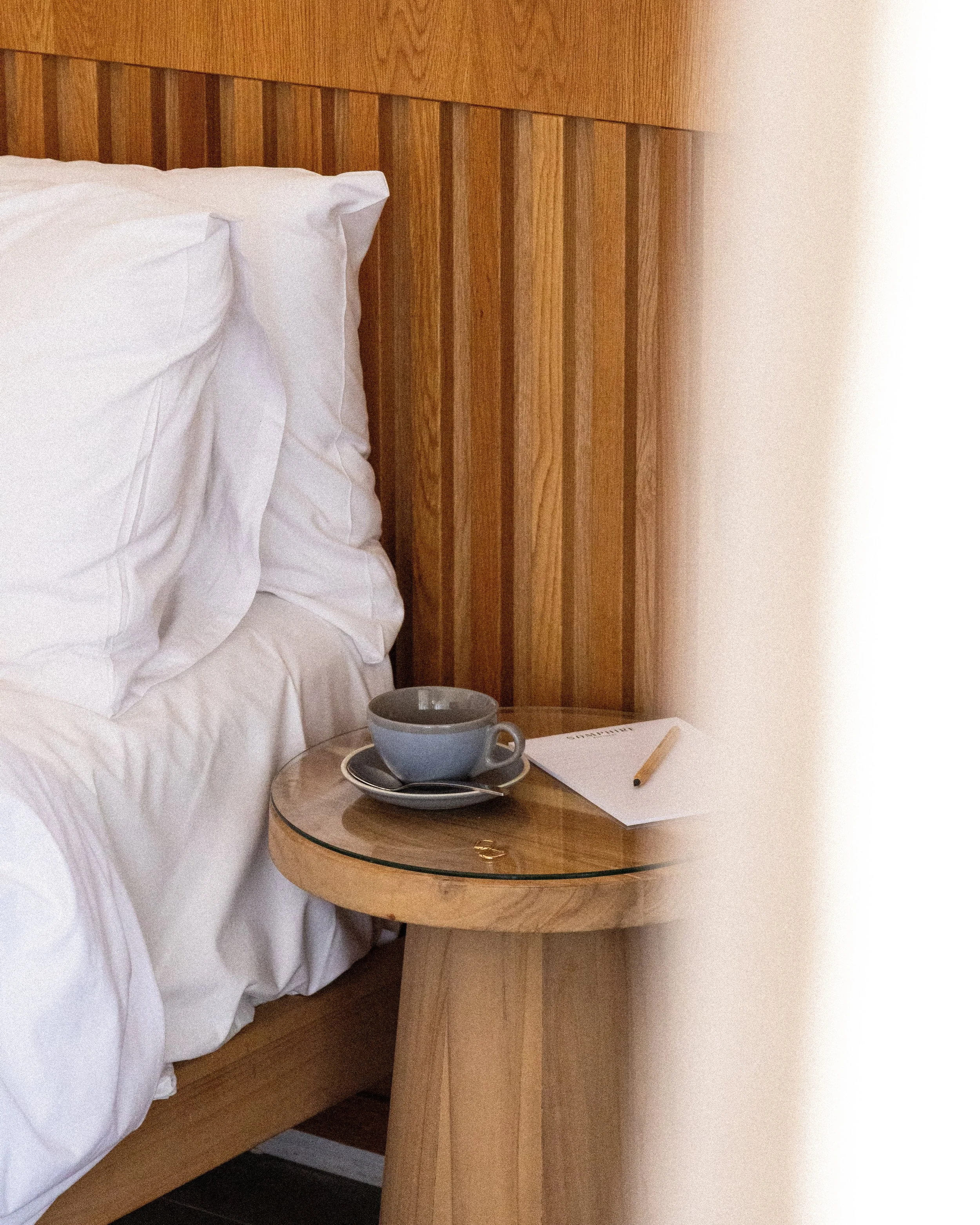 A bedroom with a wooden headboard, white pillows and bedding, a glass-top nightstand, a blue mug, a white notepad, and a pencil.