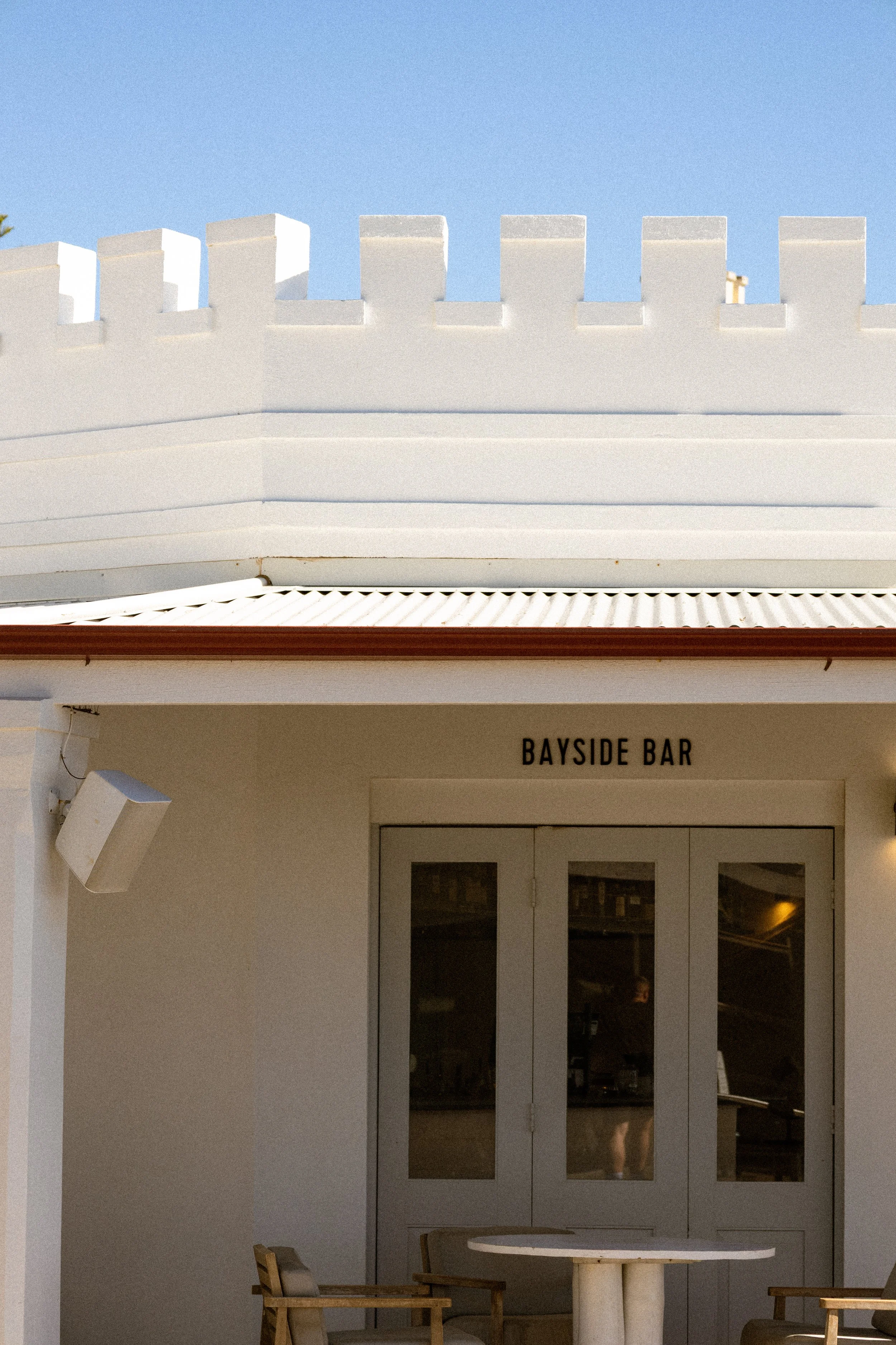 Exterior of a building with a sign that reads 'Bayside Bar', glass doors, and furniture outside, under a blue sky.