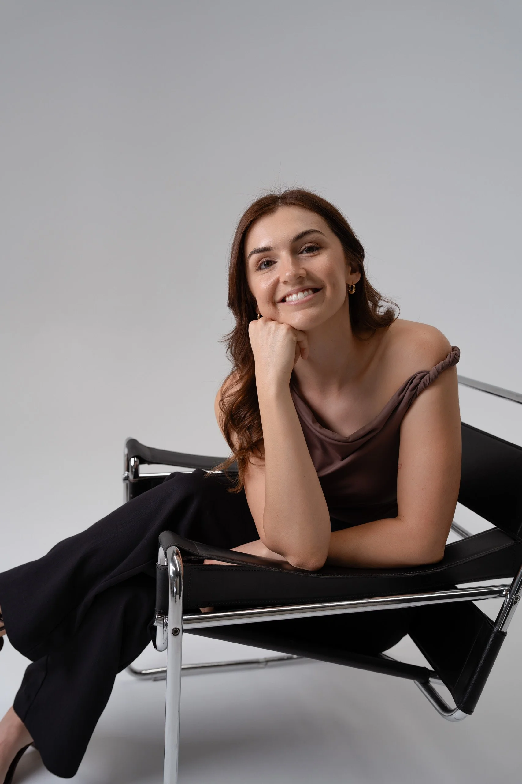 A young woman with brown hair and earrings sitting on a black lounge chair, smiling and resting her chin on her hand against a plain light gray background.