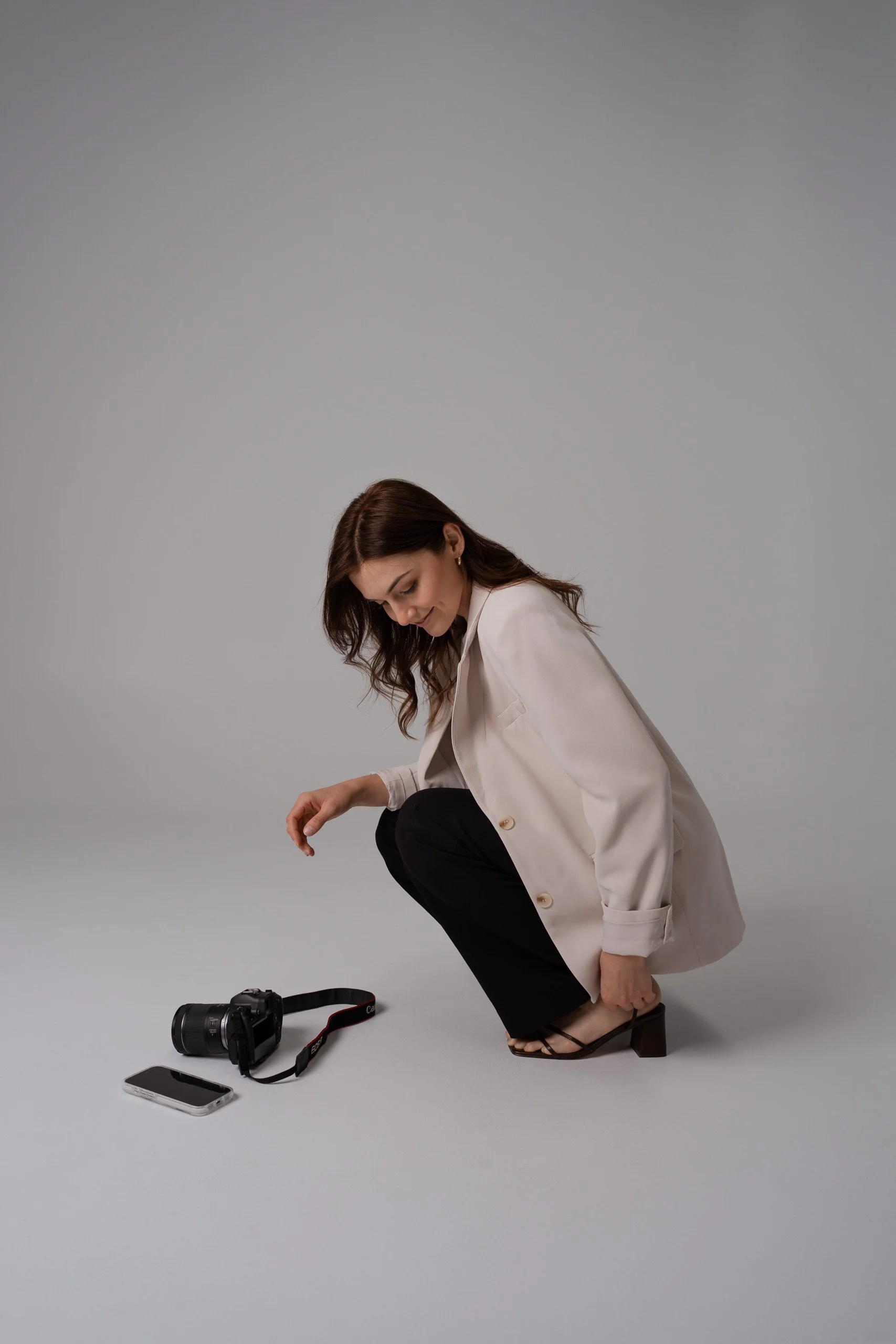 A woman with brown hair, dressed in a beige blazer and black pants, crouching down with one hand on her foot and the other on her knee, in a studio with a light gray background. Photography equipment including a camera, a phone, and a camera strap are on the floor in front of her.
