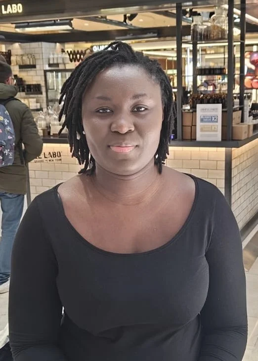 En kvinde med sorte dreadlocks iført sort langærmet top står i en butik, med en café- eller madkødsbaggrund.