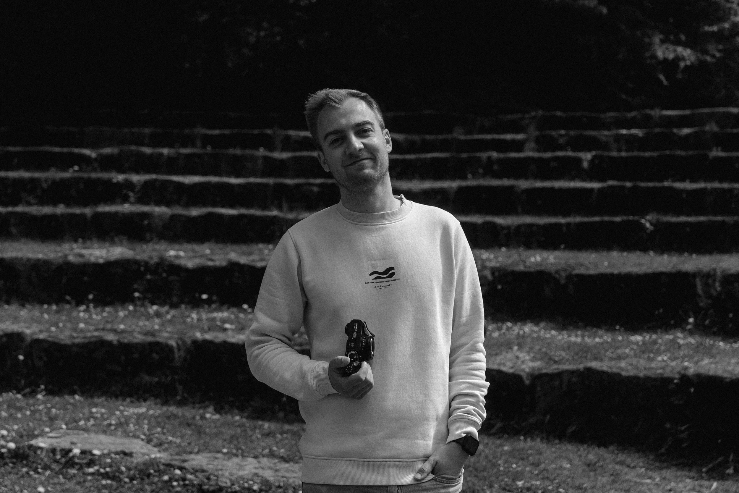 Junger Mann in hellem Sweatshirt hält eine Kamera in der Hand, im Hintergrund treppenartige Steigung aus Steinen, nächtliche Umgebung.