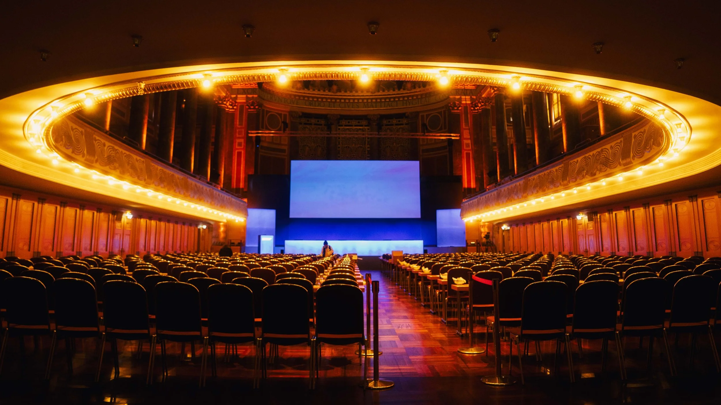 Ein leeres großes Konzerthalle mit vielen Sitzplätzen, einer Bühne mit großen Bildschirmen und moderner Lichtgestaltung.