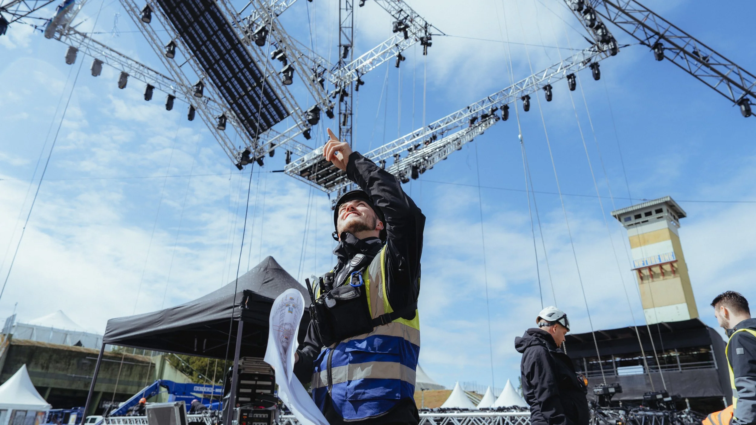 Mann am Festival- oder Konzertgelände, der in der Hand eine Plan hält, während unter einem großen Bühnen- und Lichterrahmen im Freien ein Team arbeitet. Der Himmel ist leicht bewölkt.