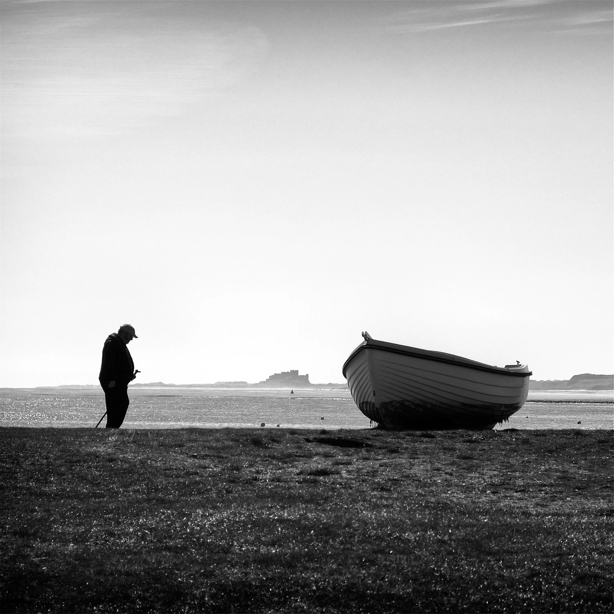 Silhouette of a person standing near an old fishing boat, with the sea and a distant castle in the background, under a clear sky.