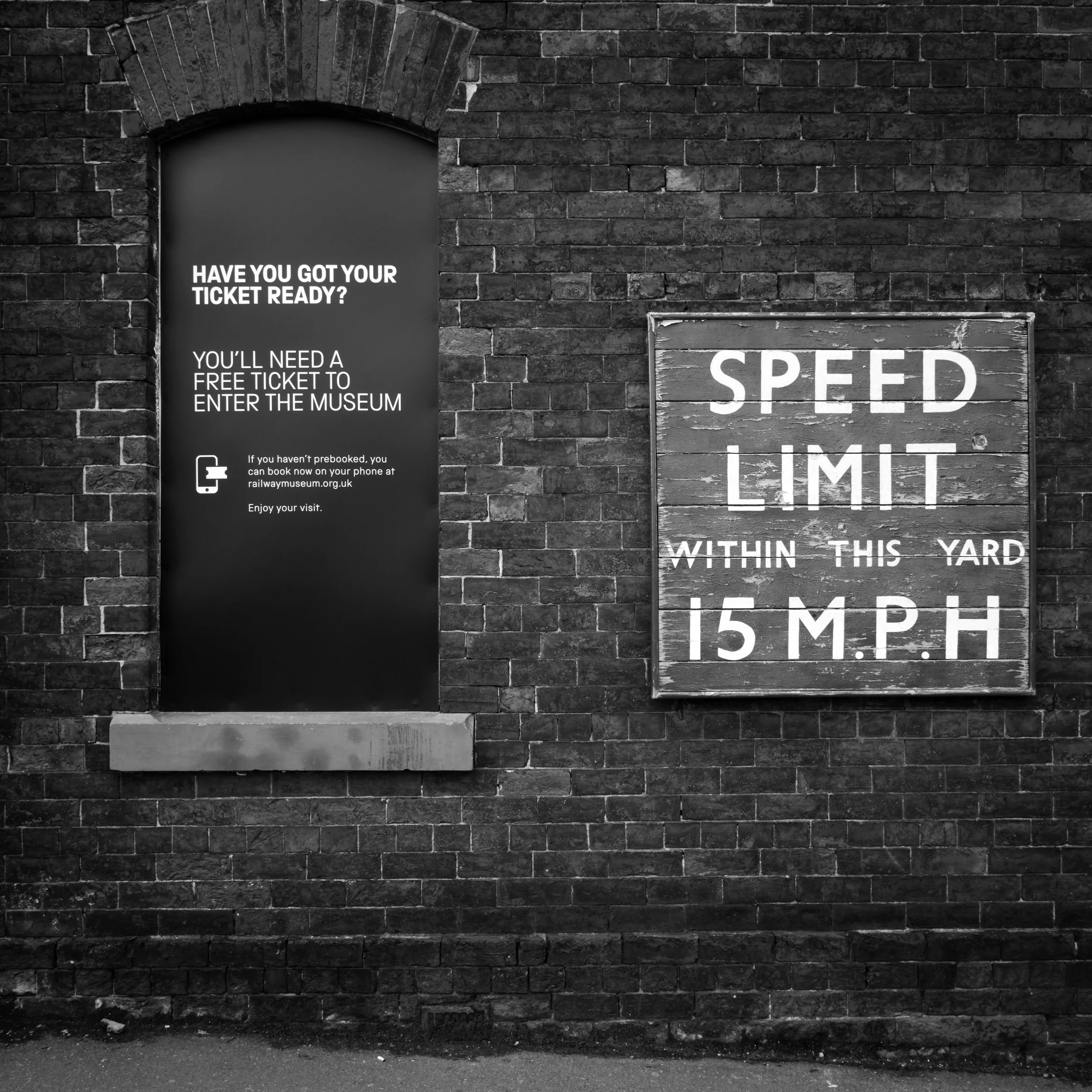 Black brick wall with a small arched doorway and a signboard that reads 'Speed Limit 5 MPH', located within a yard.