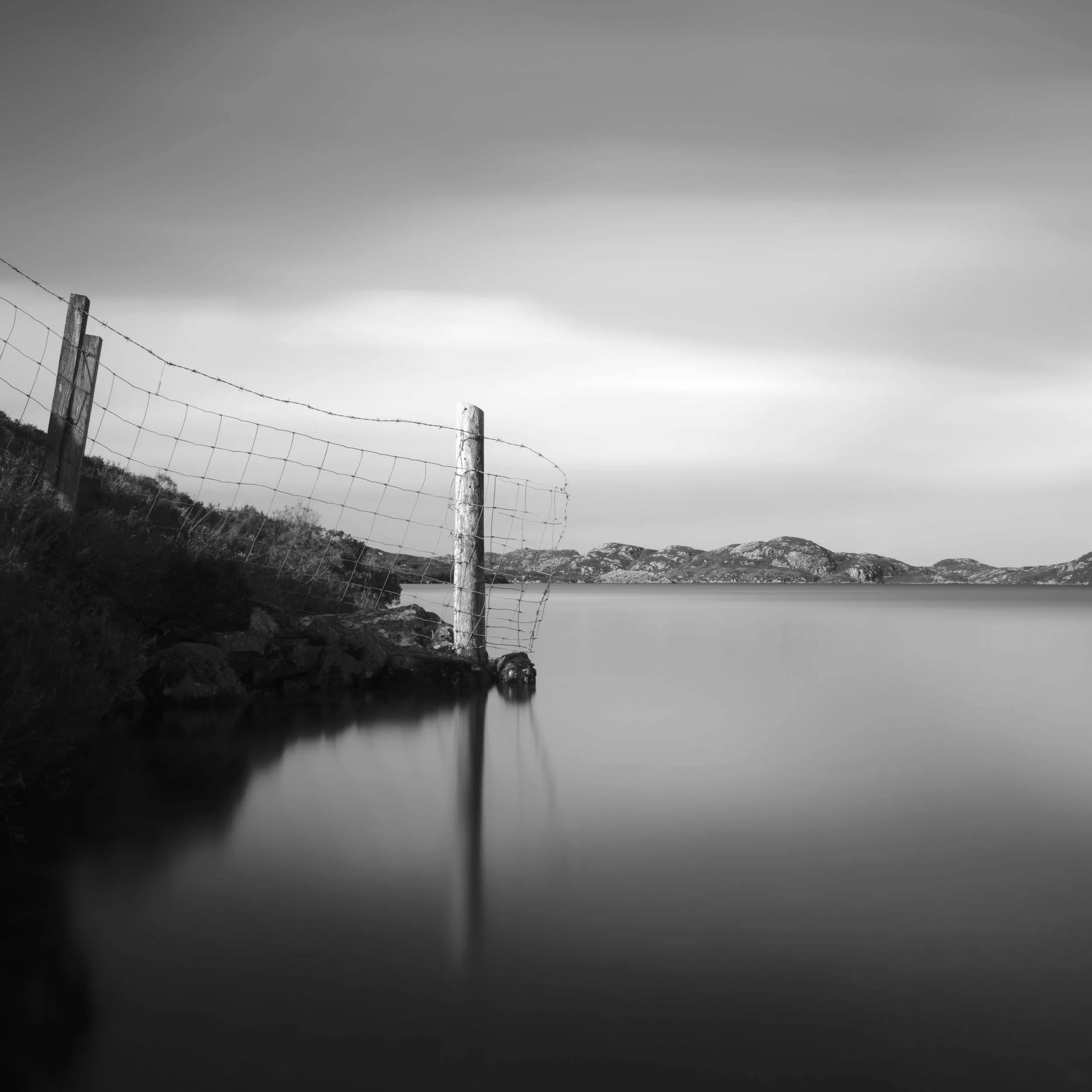 Black and white long exposure landscape photo of a loch surrounded by hills or mountains with a cloudy sky, featuring a reflection of a wire fence with wooden posts in the foreground.