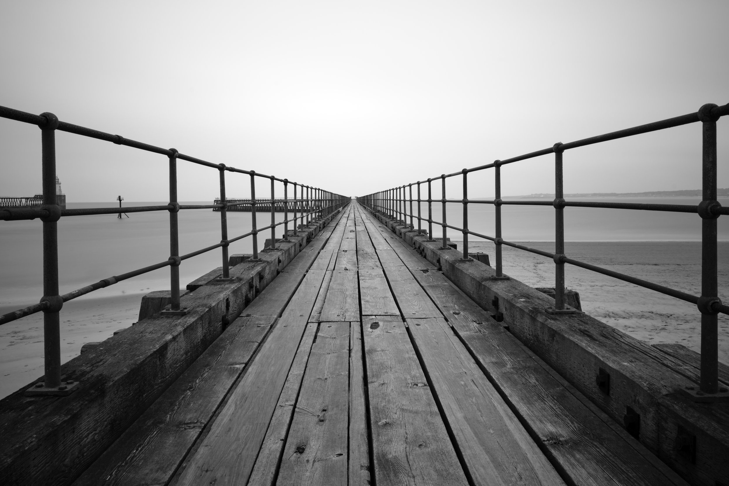 Black and white photo of a long wooden pier extending into the sea, with metal railings on both sides, leading to a distant lighthouse.