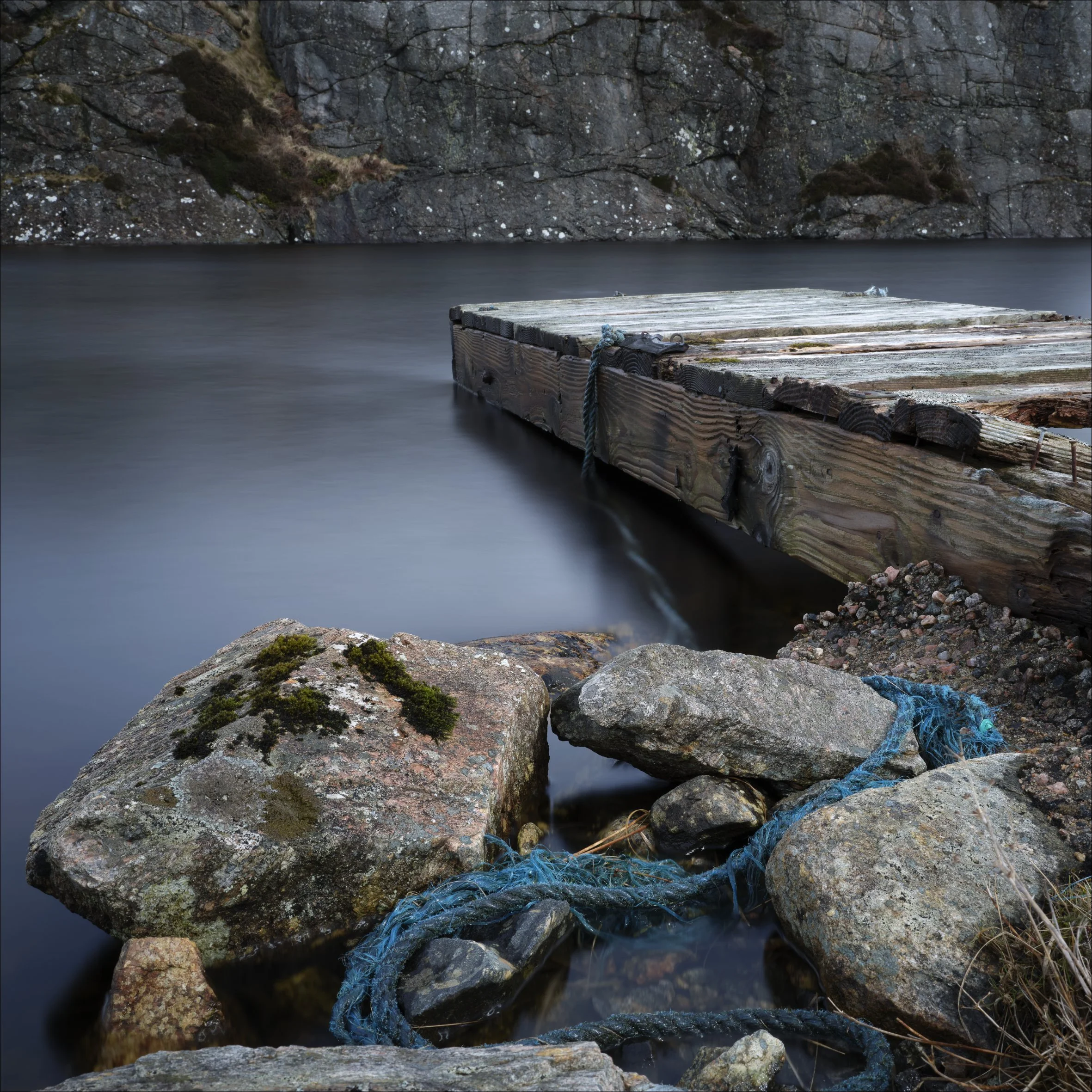 A wooden dock extending into a calm body of water with rocky and moss-covered terrain in the background.