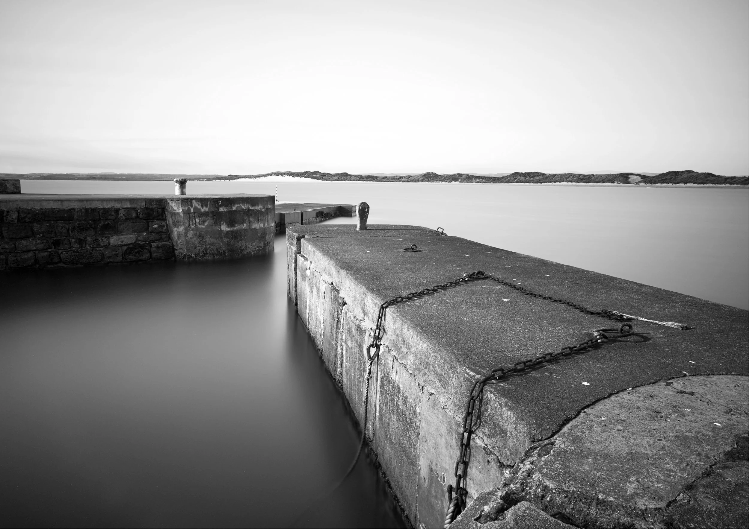 A black and white photo of a two stone piers extending into the ocean. In the background, there are dunes on the horizon along the coastline under a clear sky.