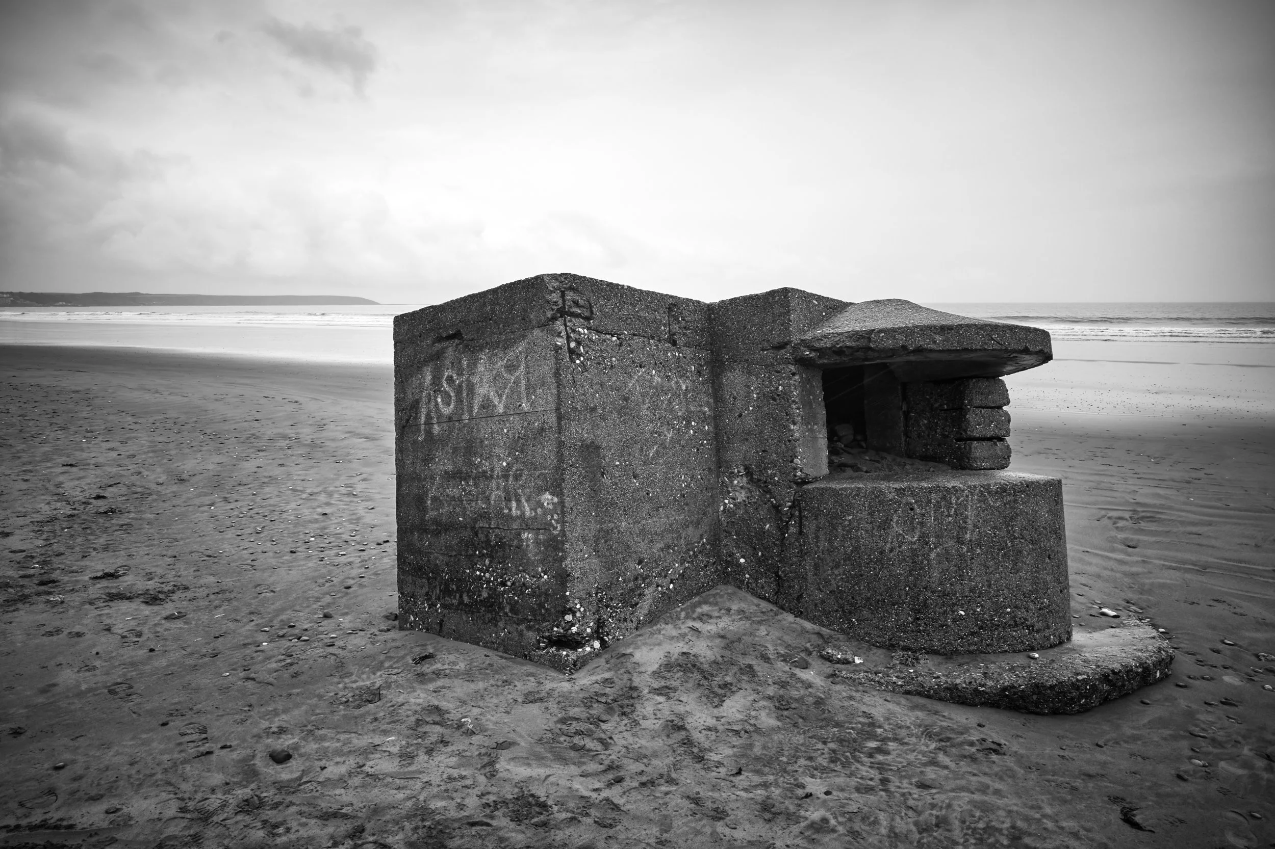 Black and white photo of a concrete structure on a sandy beach, with the ocean and cloudy sky in the background.