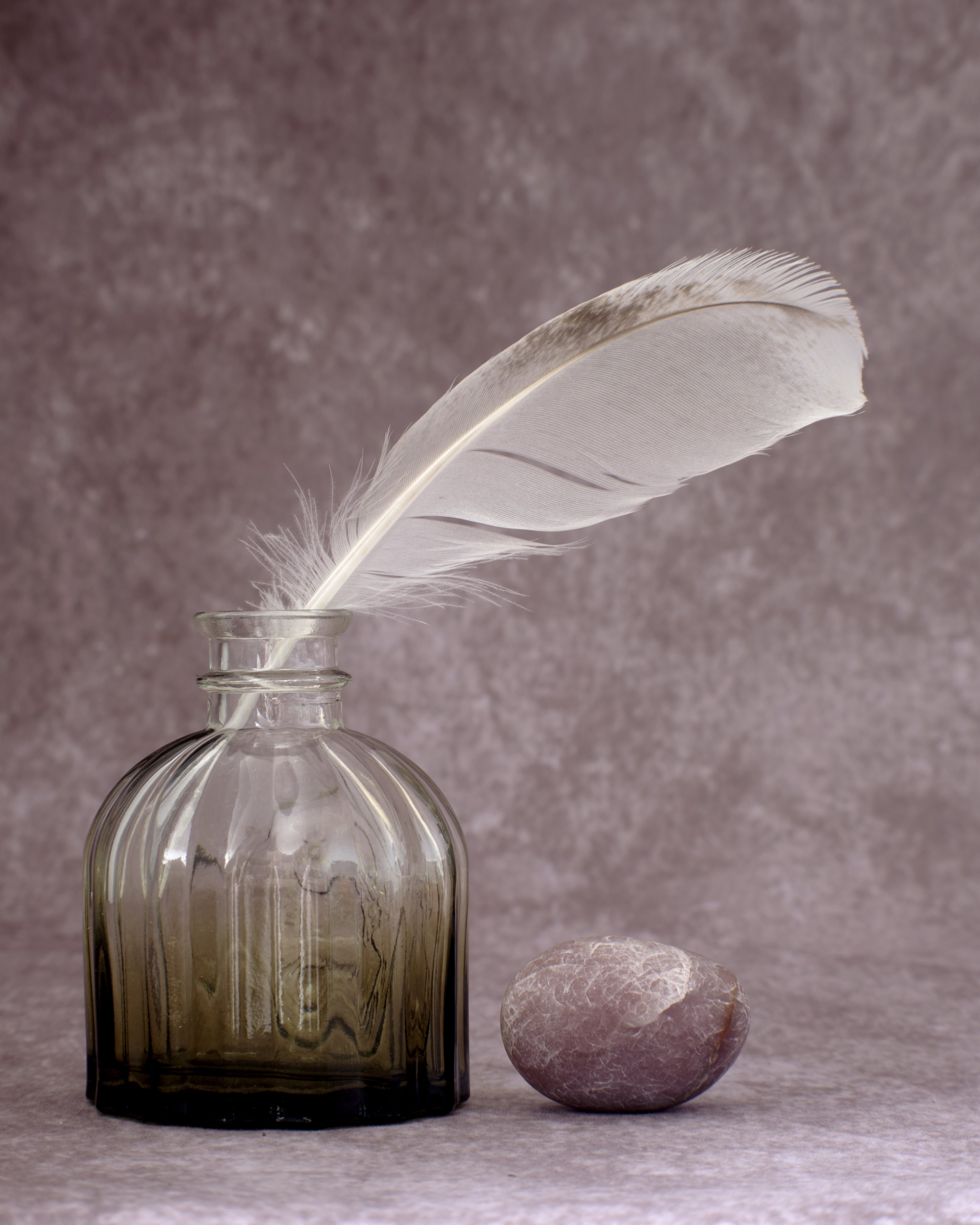 A clear glass oil container with vertical ridges, with a white feather standing inside it, a pinkish-white stone on the surface in front of the container, and a textured pink background.