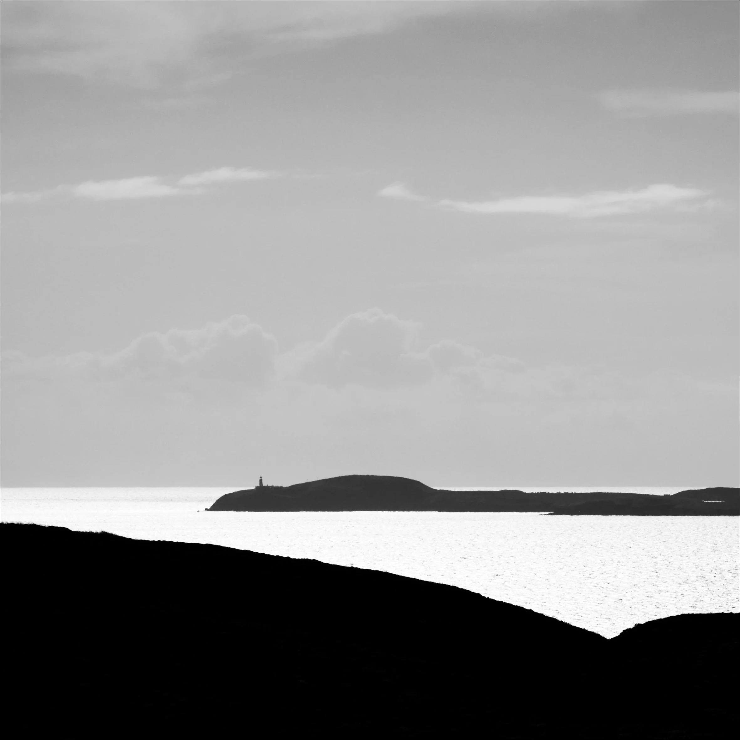 Silhouette of a lighthouse on a island headland, with the sea surrounding it. The sky is cloudy and the scene is in atmospheric black and white.