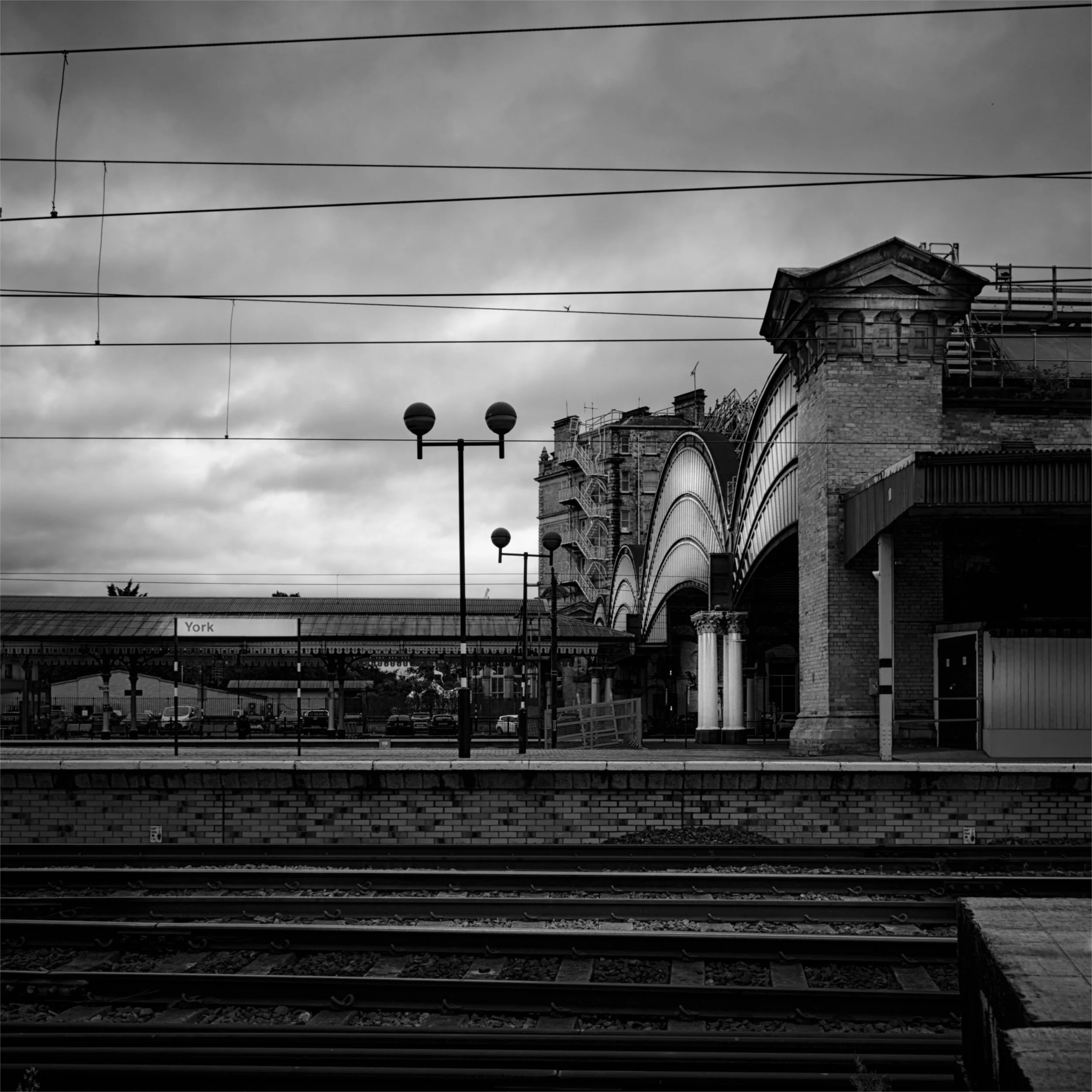 Black and white photo of a train platform with overhead wires, a clock, and an ornate building with scaffolding. The sky is cloudy.