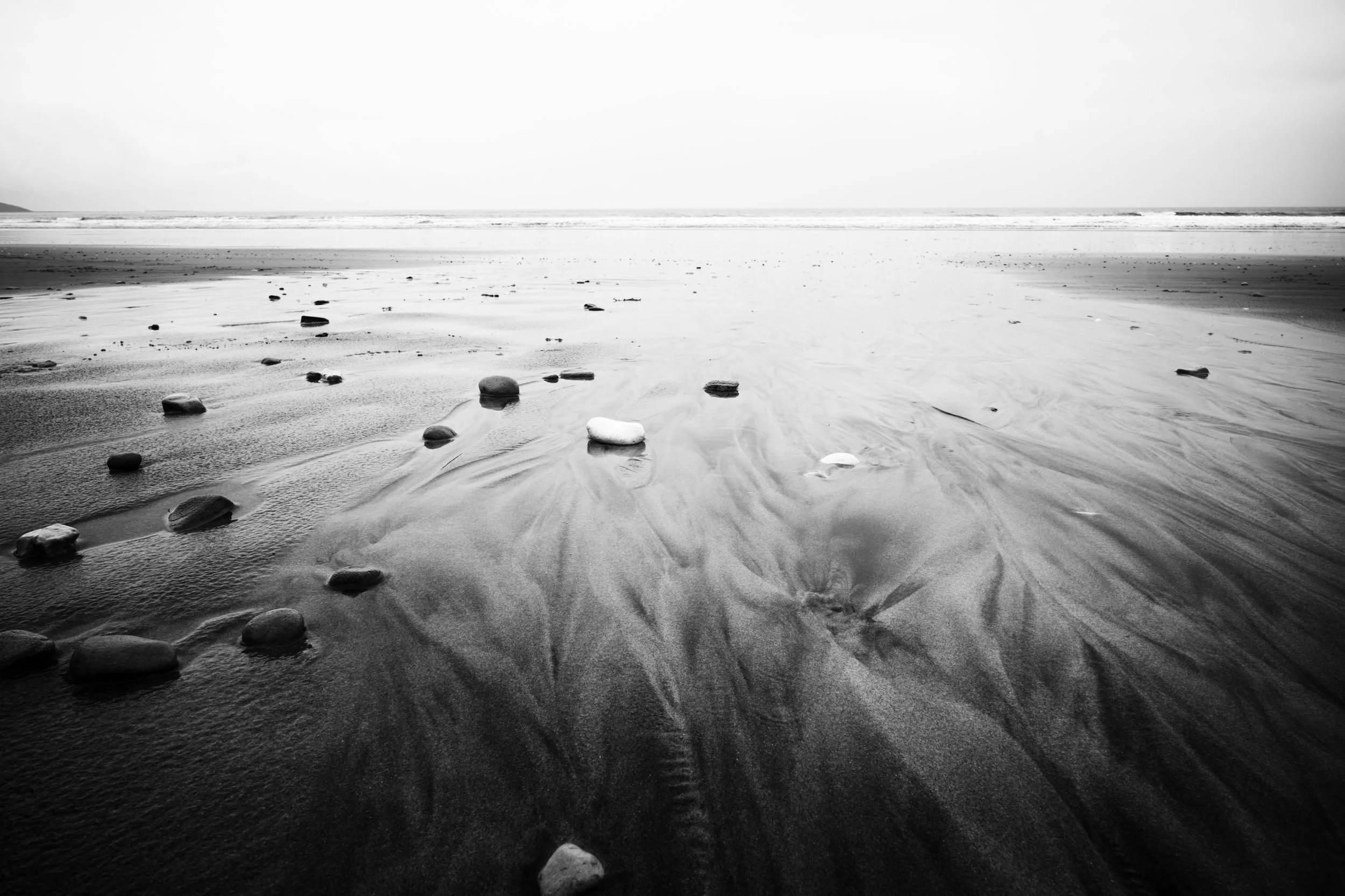 Black and white photograph of a sandy beach with rocks scattered across the sand, sea in the background, overcast sky.