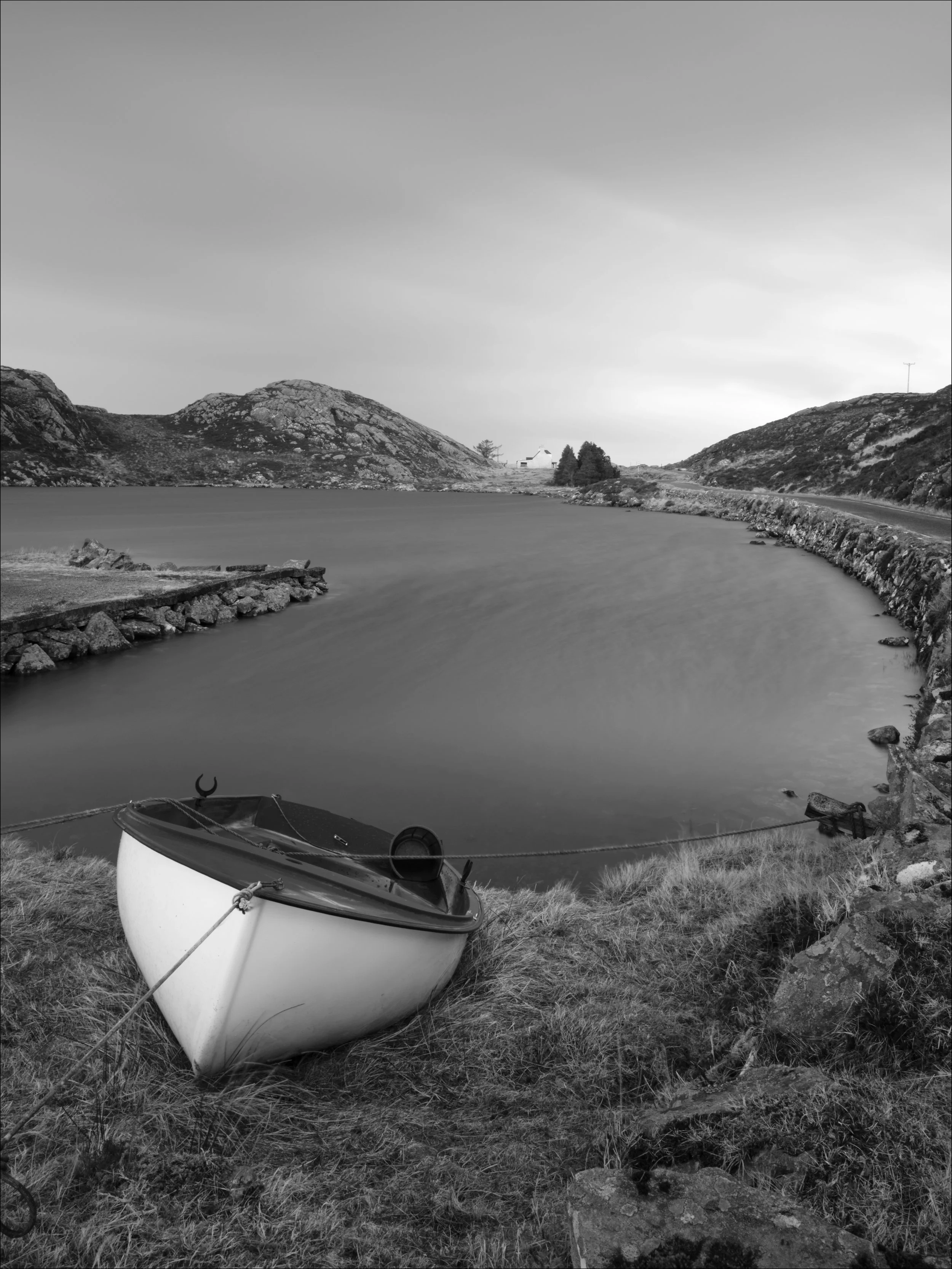 A long exposure black and white photo of a small boat resting on grassy shore with rocks, overlooking a calm loch surrounded by hilly terrain, under a cloudy sky.