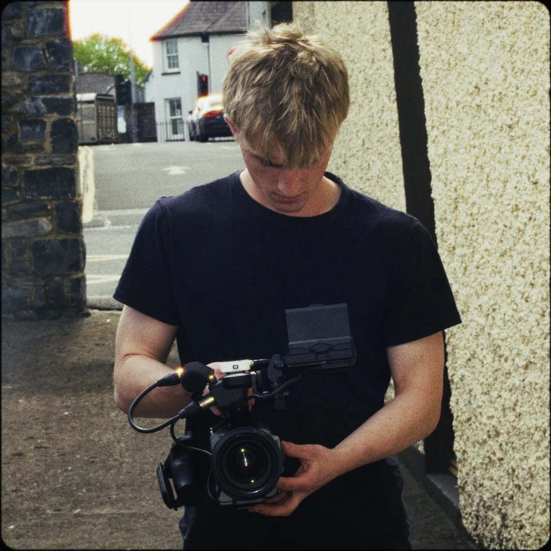 Young man with blond hair holding a professional camera and looking down, standing outdoors near a stone wall with a street and houses in the background.