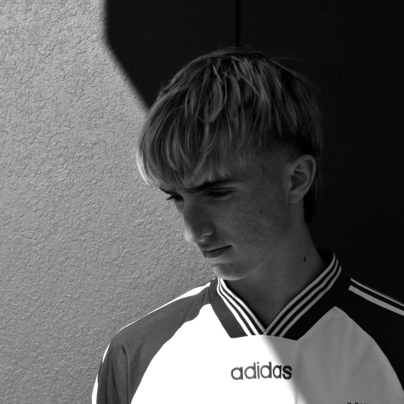 A young person with short, light-colored hair looking down, photographed in black and white against a textured wall, wearing an Adidas sports jersey.