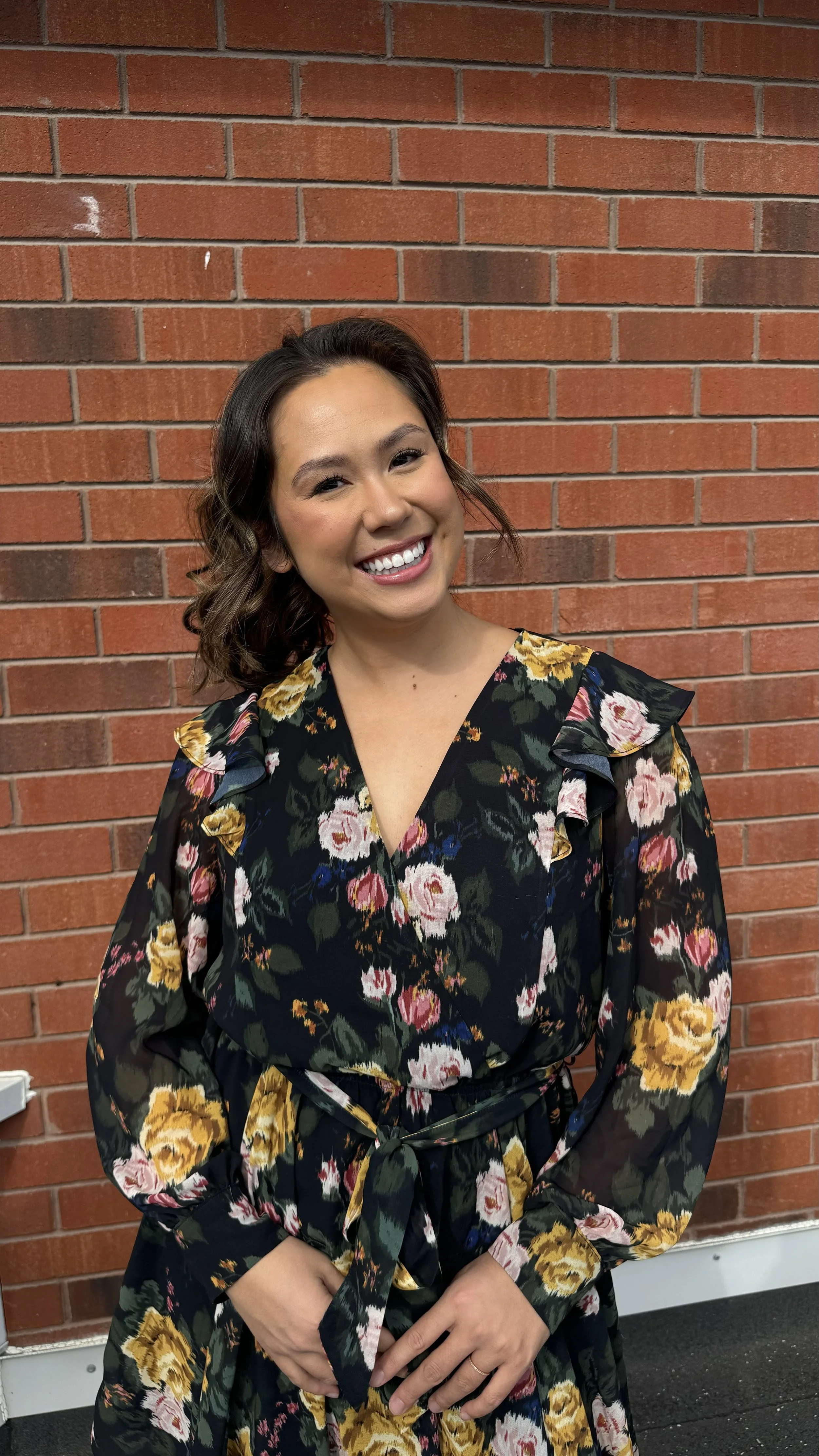 A smiling woman with shoulder-length wavy hair wearing a black floral dress with pink, yellow, green, and white flowers, standing in front of a red brick wall.