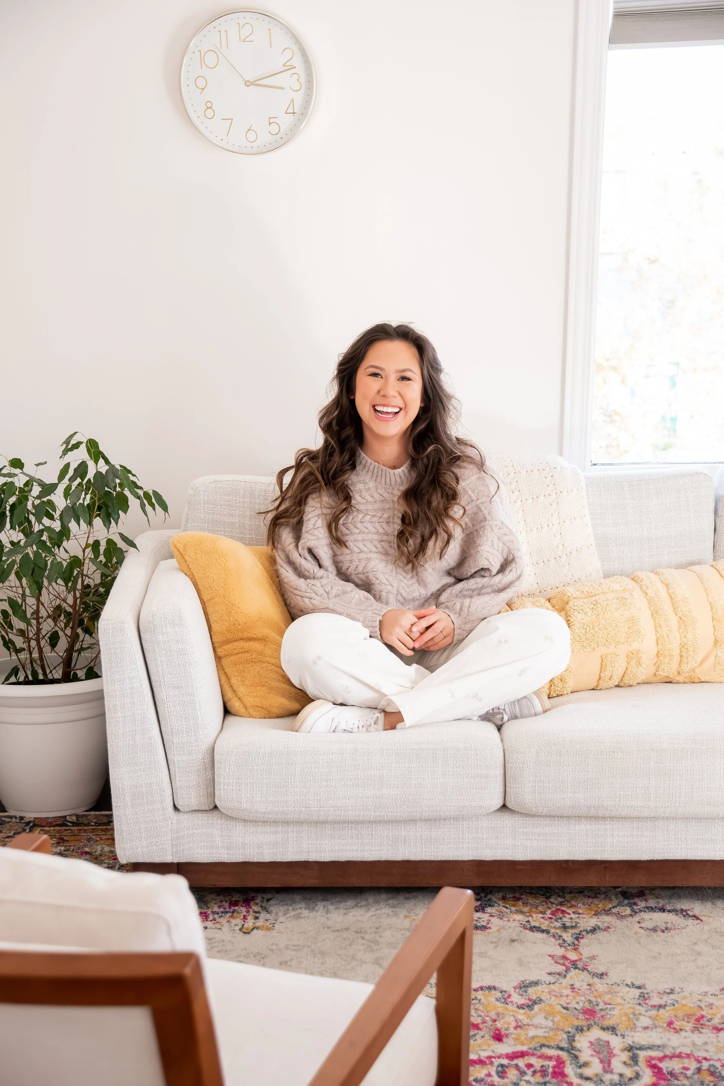 A woman sitting cross-legged on a light gray sofa, smiling at the camera. She is wearing a beige knitted sweater and white pants. Behind her, there are yellow and beige throw pillows. To the left, a large potted plant, and to the right, a window with sunlight coming in. A white clock showing 2:13 is on the wall above.