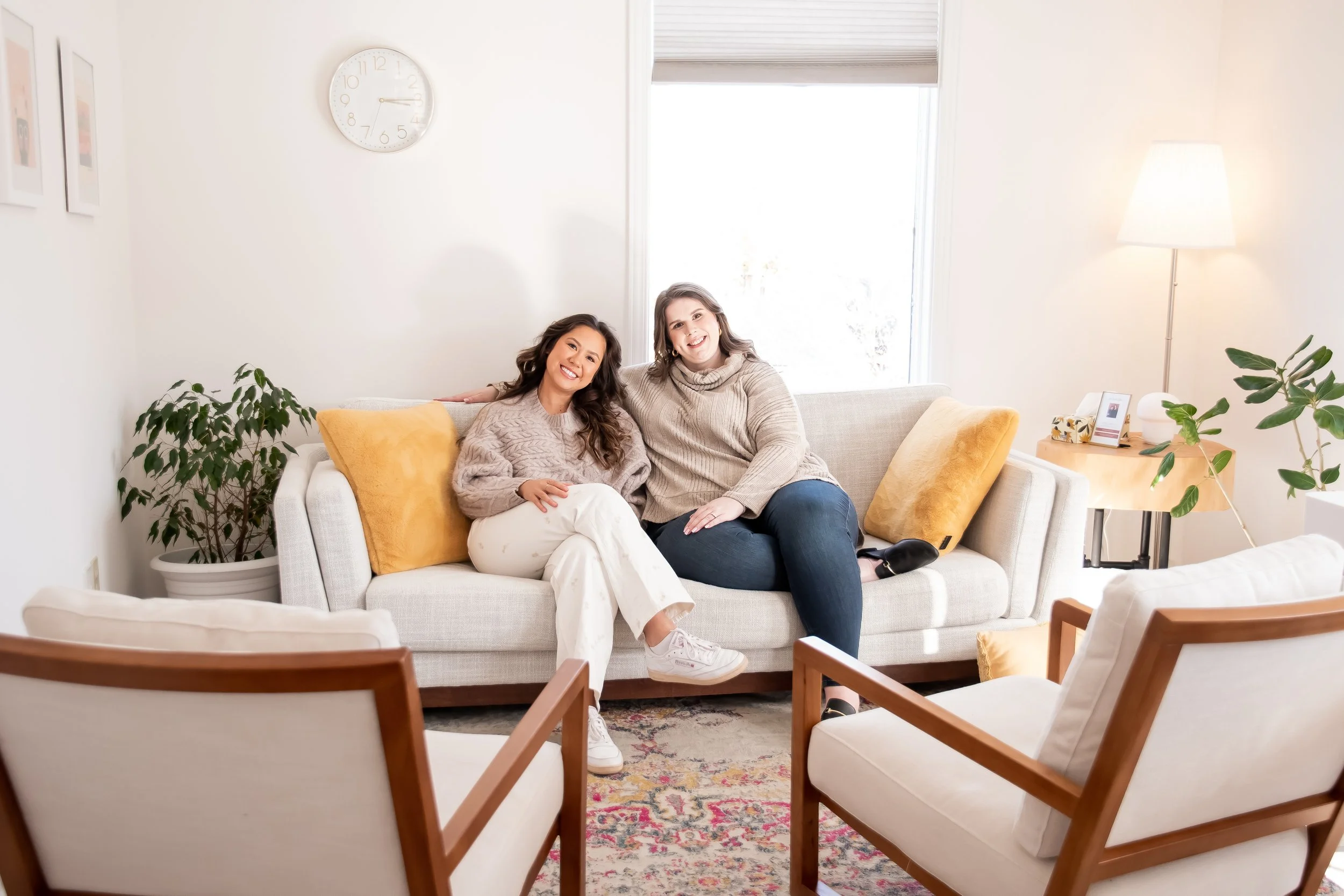 Two women sitting on a beige sofa in a bright living room, smiling and posing for the photo. There are yellow pillows on the sofa, a potted plant, a wooden side table with a lamp, a framed picture, and a large window in the background.
