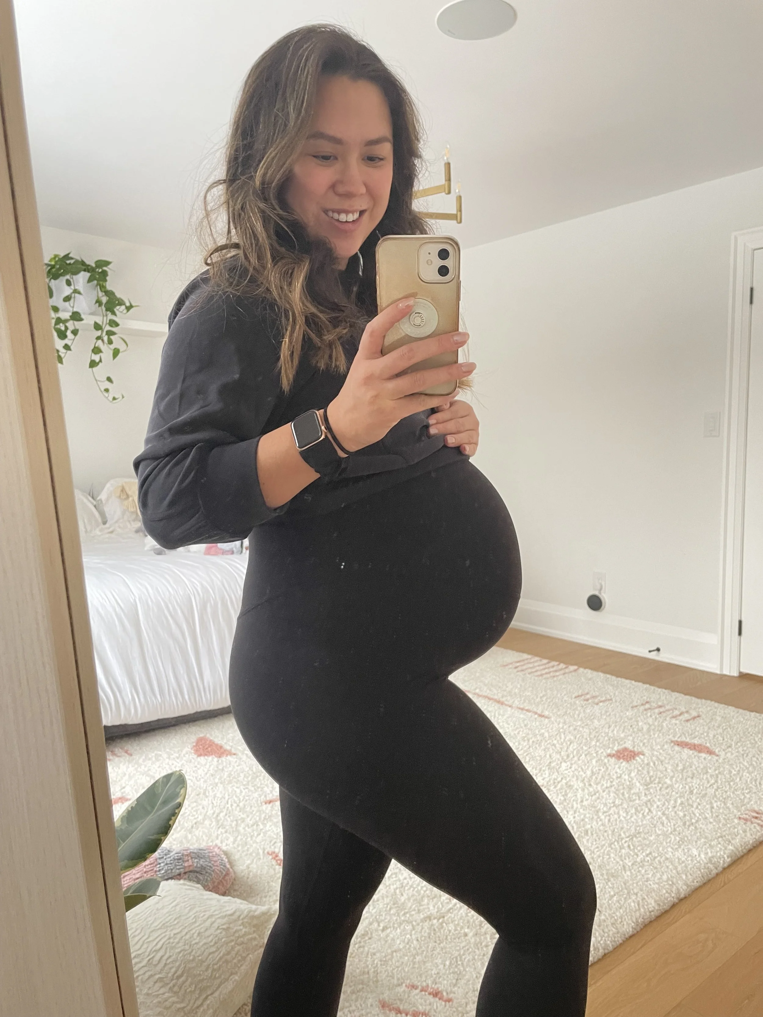 A pregnant woman taking a selfie in front of a mirror, smiling, wearing a black long sleeve top and black leggings, standing in a bedroom.