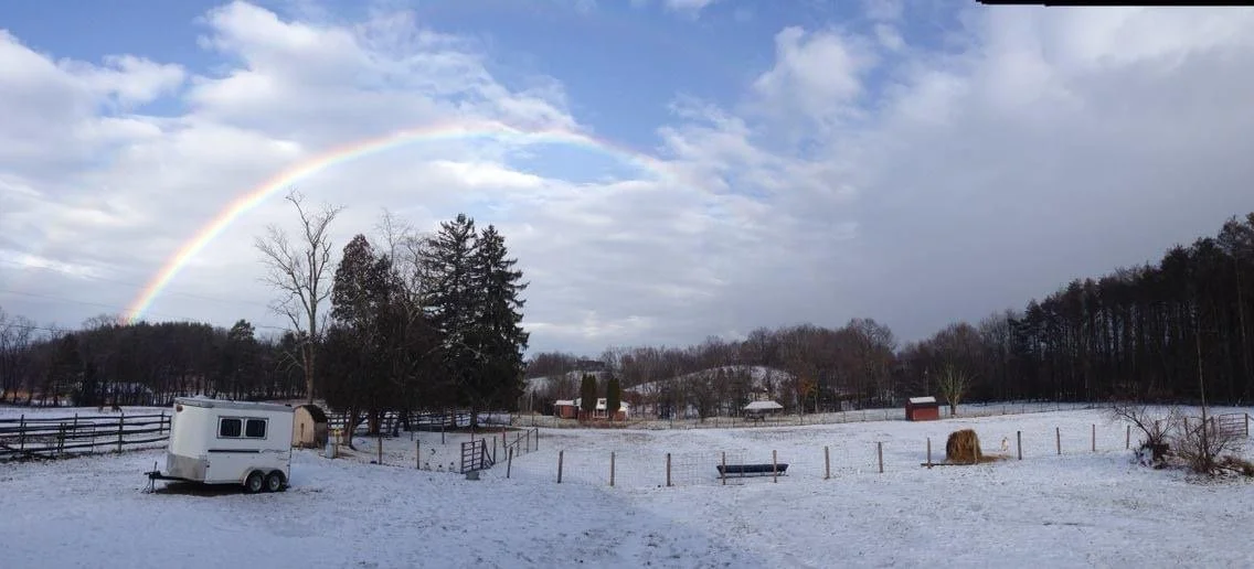 Snow-covered farm landscape with trees, a rainbow in the cloudy sky, and a small white trailer in the foreground.