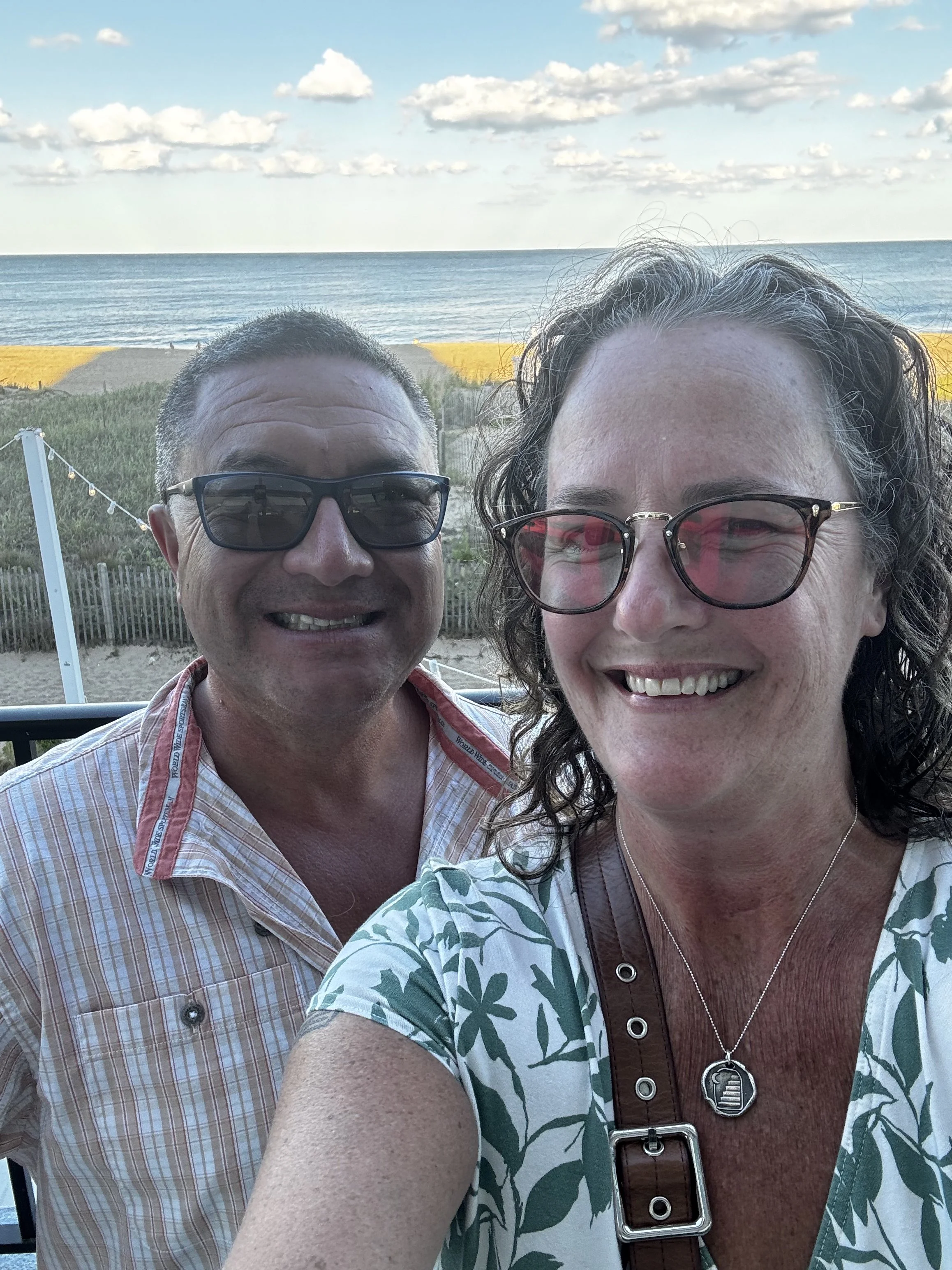 A smiling couple taking a selfie with the beach ocean and sky in the background. The woman has curly hair, glasses, and is wearing a patterned shirt, while the man has short hair, glasses, and is dressed in a checkered shirt.